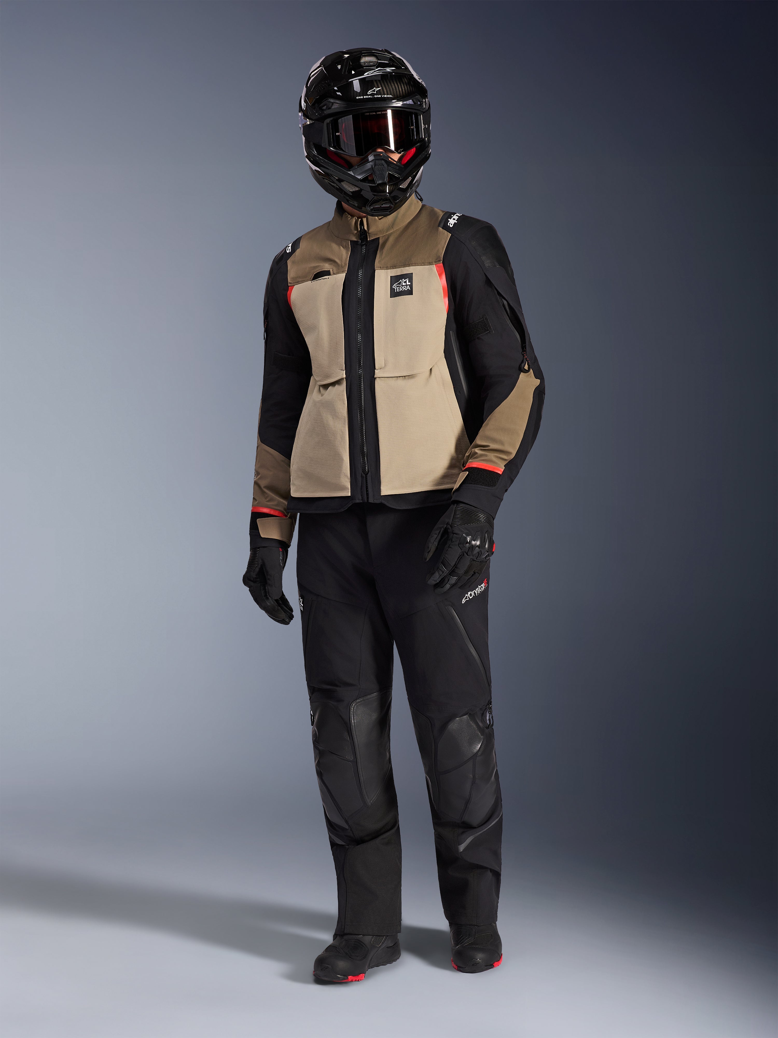 Chaqueta Alpinestars Andes Pro Drystar® XF en Walnut Aluminum, chaqueta de motociclismo de aventura combinada con pantalones negros a juego, casco adventure negro con gafas y botas negras, con paneles en color arena y negro con detalles en rojo.