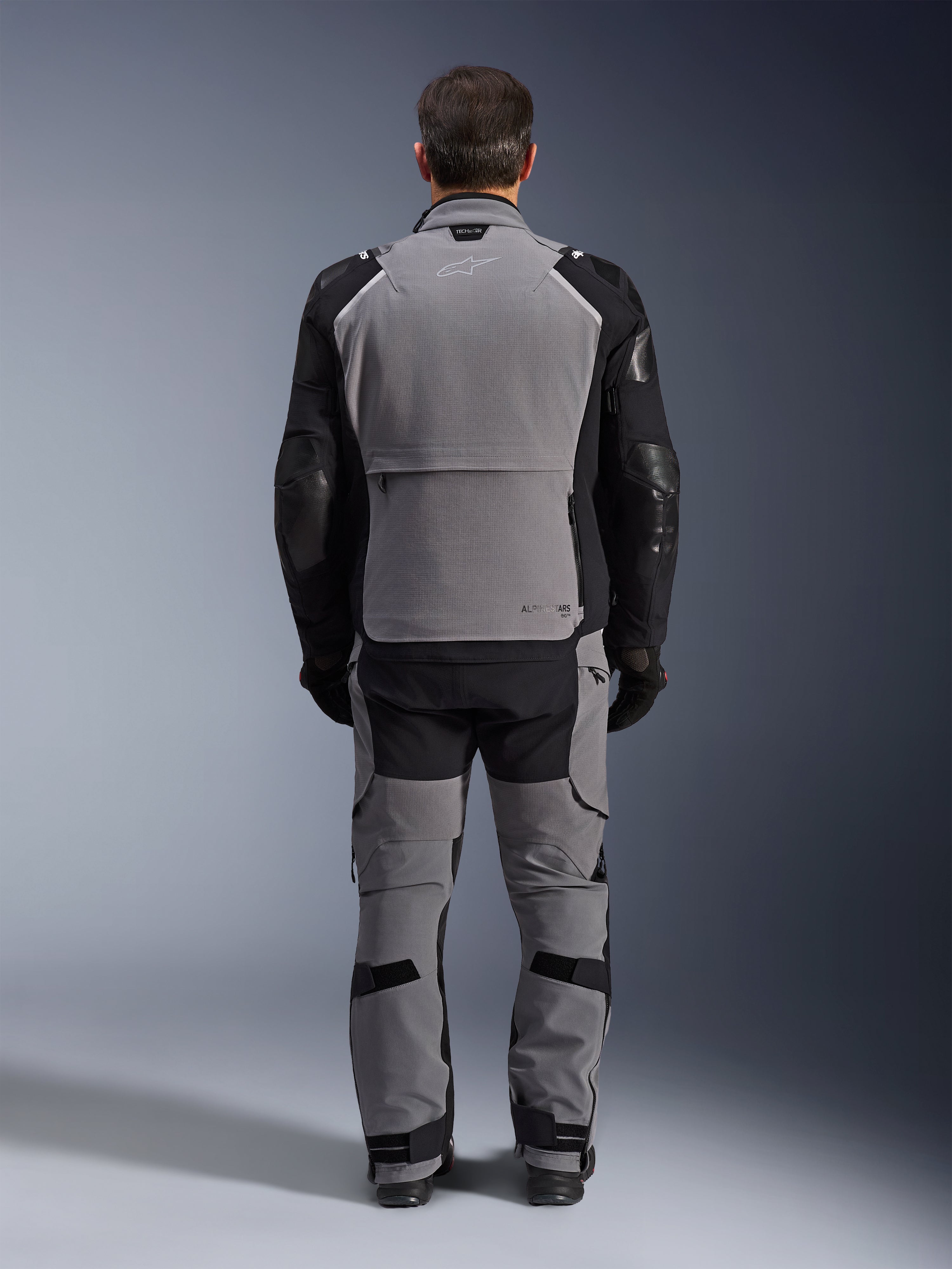 Chaqueta Alpinestars Andes Pro Drystar® XF, Gris Oscuro Negro, vista trasera de un piloto con pantalones a juego, chaqueta de motociclismo para adventure touring con un gran panel gris en la espalda, mangas articuladas negras con protecciones y un compartimento de almacenamiento trasero