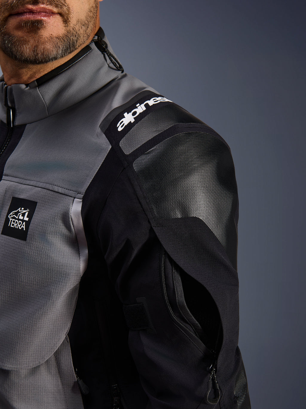 Chaqueta Alpinestars Andes Pro Drystar® XF, chaqueta de moto de aventura, gris oscuro y negro, vista de primer plano del hombro reforzado con la marca Alpinestars, parche con el logotipo 'TERRA' y ventilación con cremallera en la manga sobre una capa de tejido técnico.