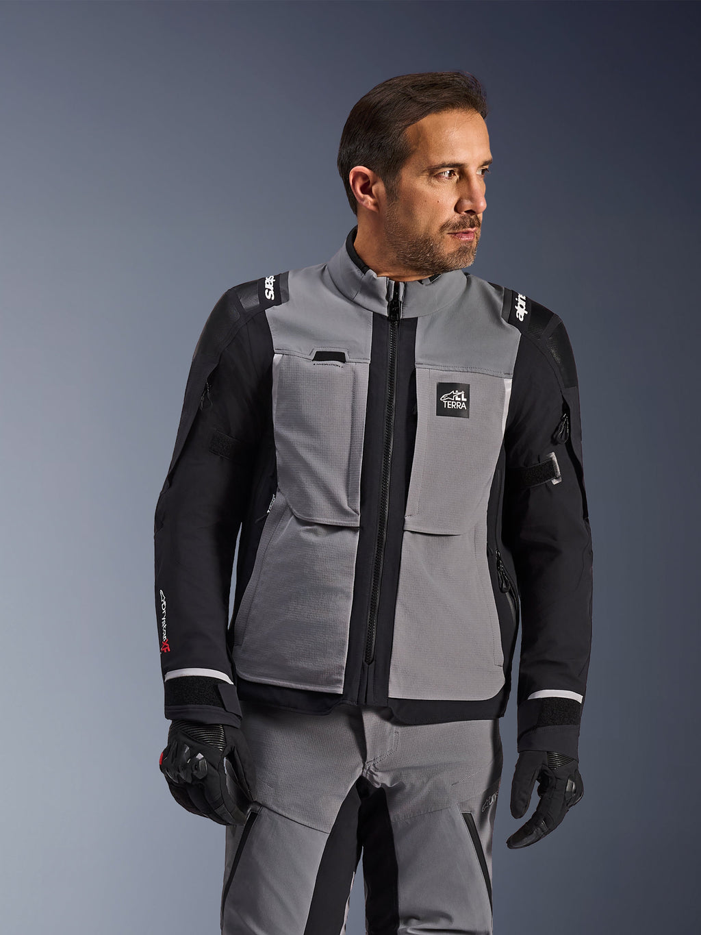 Chaqueta Alpinestars Andes Pro Drystar® XF en gris oscuro y negro, equipamiento de aventura para motociclistas, usada por un hombre con pantalones grises coordinados y guantes negros, con capa laminada de 3 capas, bolsillos utilitarios y logotipo 'Terra' en el pecho