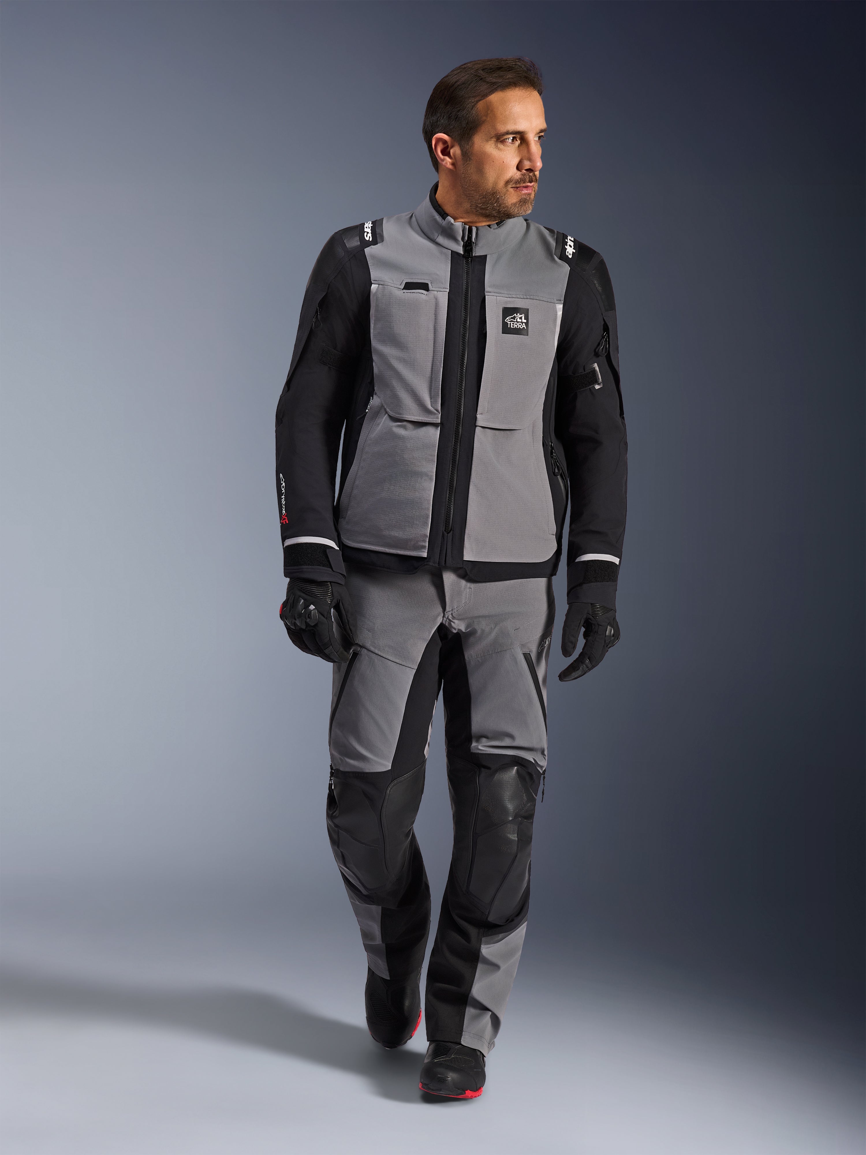 Modelo masculino vistiendo la chaqueta Alpinestars Andes Pro Drystar® XF en gris oscuro y negro, traje de motociclismo de aventura con pantalones y guantes a juego, con una capa de tejido técnico y marca 'All Terra', equipamiento completo para touring de aventura de larga distancia