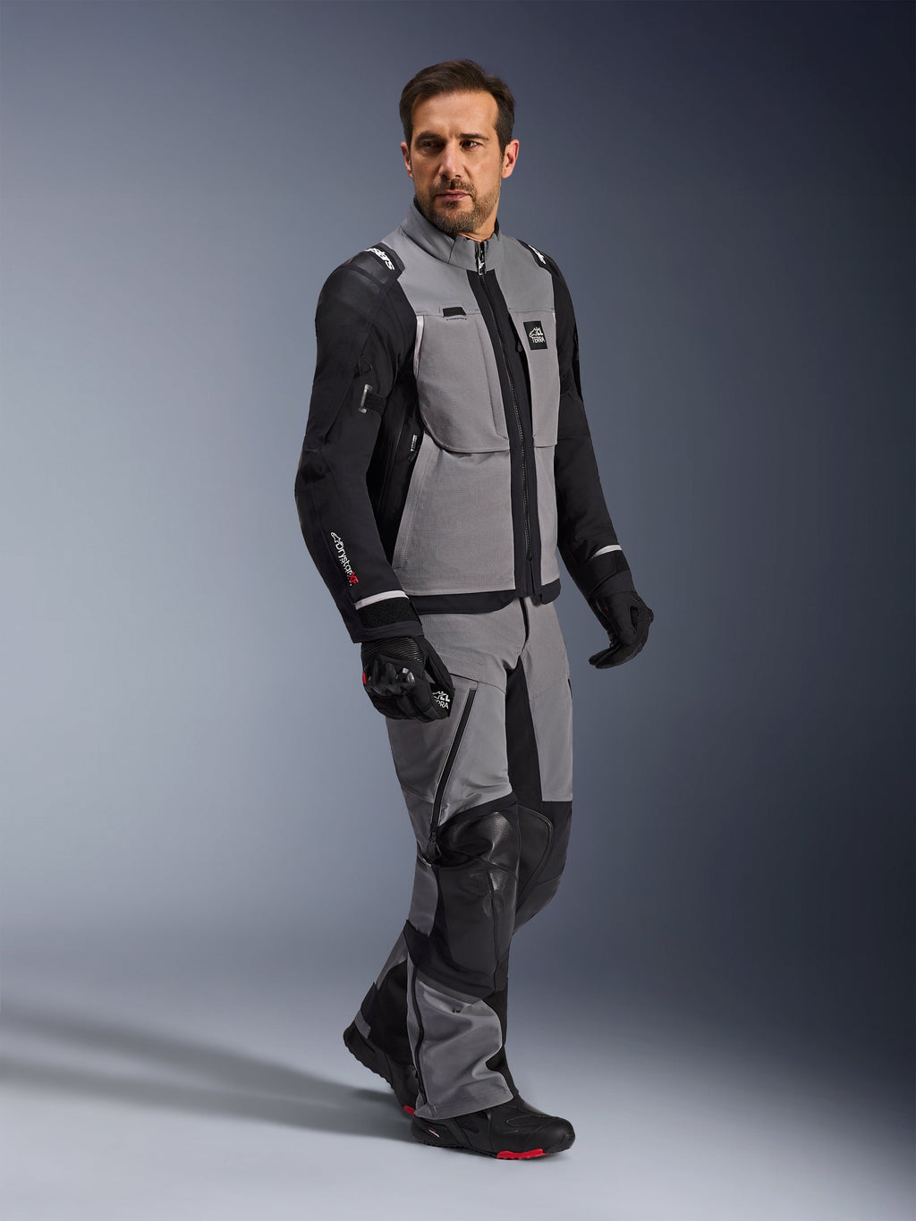 Chaqueta Alpinestars Andes Pro Drystar® XF, chaqueta de motociclismo de aventura, gris oscuro y negro, lucida por un modelo con pantalones textiles a juego y guantes negros, con diseño multibolsillo, capa técnica laminada de 3 capas y ajuste articulado para viajes de aventura de larga distancia.