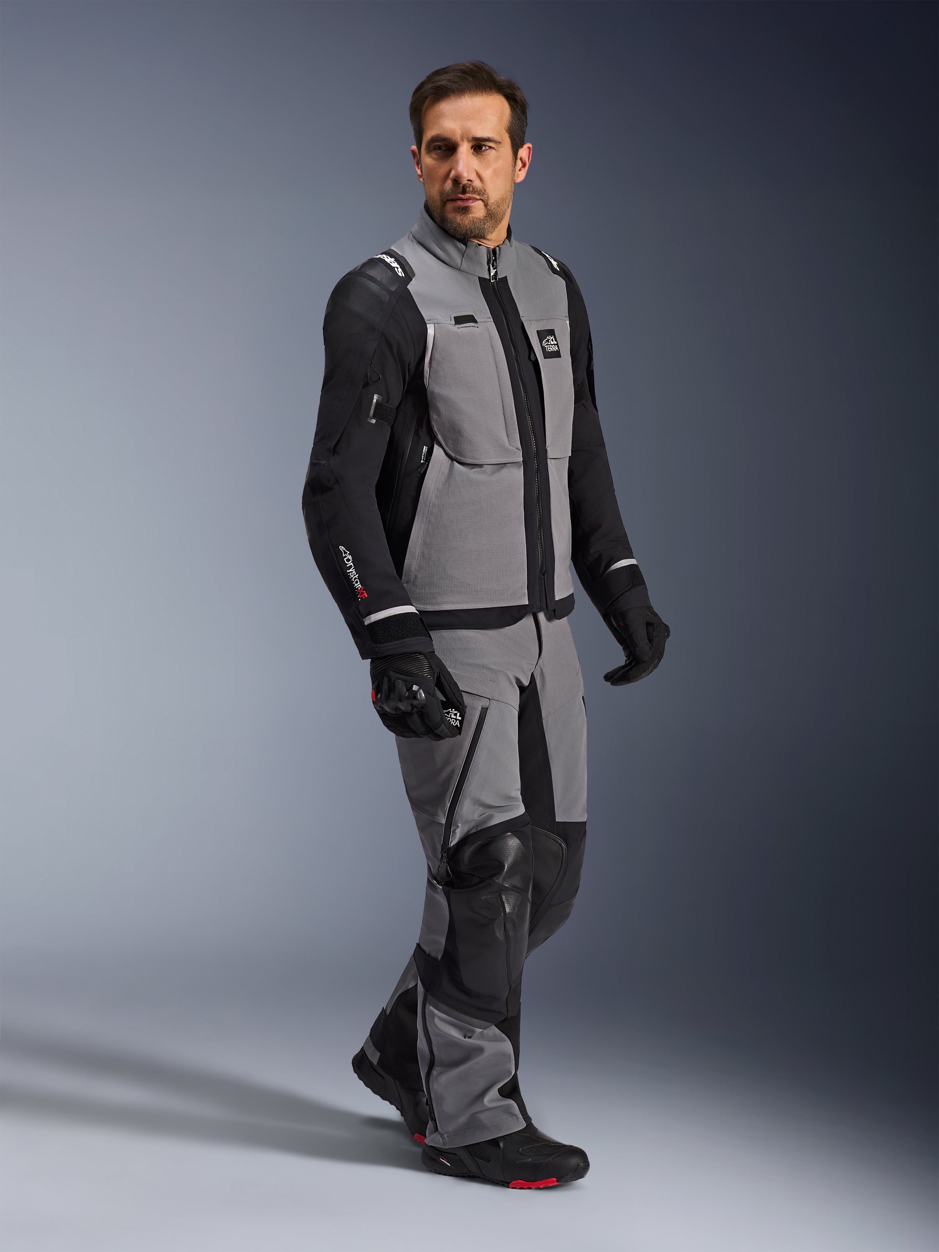 Chaqueta Alpinestars Andes Pro Drystar® XF, chaqueta de motociclismo de aventura, gris oscuro y negro, lucida por un modelo con pantalones textiles a juego y guantes negros, con diseño multibolsillo, capa técnica laminada de 3 capas y ajuste articulado para viajes de aventura de larga distancia.
