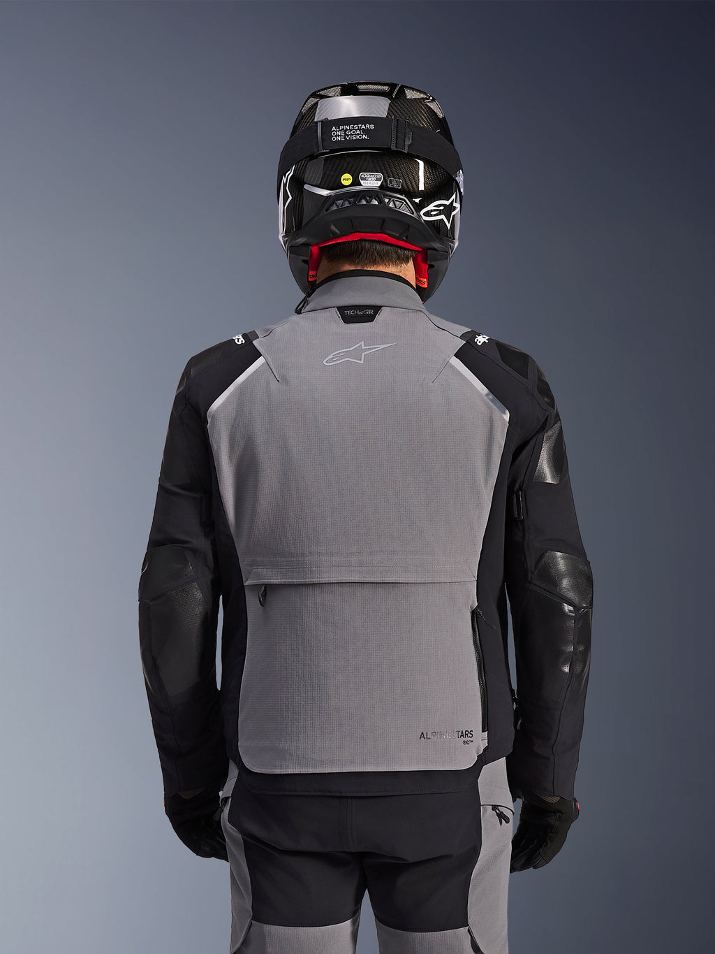Chaqueta Alpinestars Andes Pro Drystar® XF, gris oscuro y negro, vista trasera que muestra el gran bolsillo de almacenamiento posterior y la marca Tech-Air, usada por un motorista con casco negro y pantalones de conducción a juego, equipamiento profesional para touring de aventura
