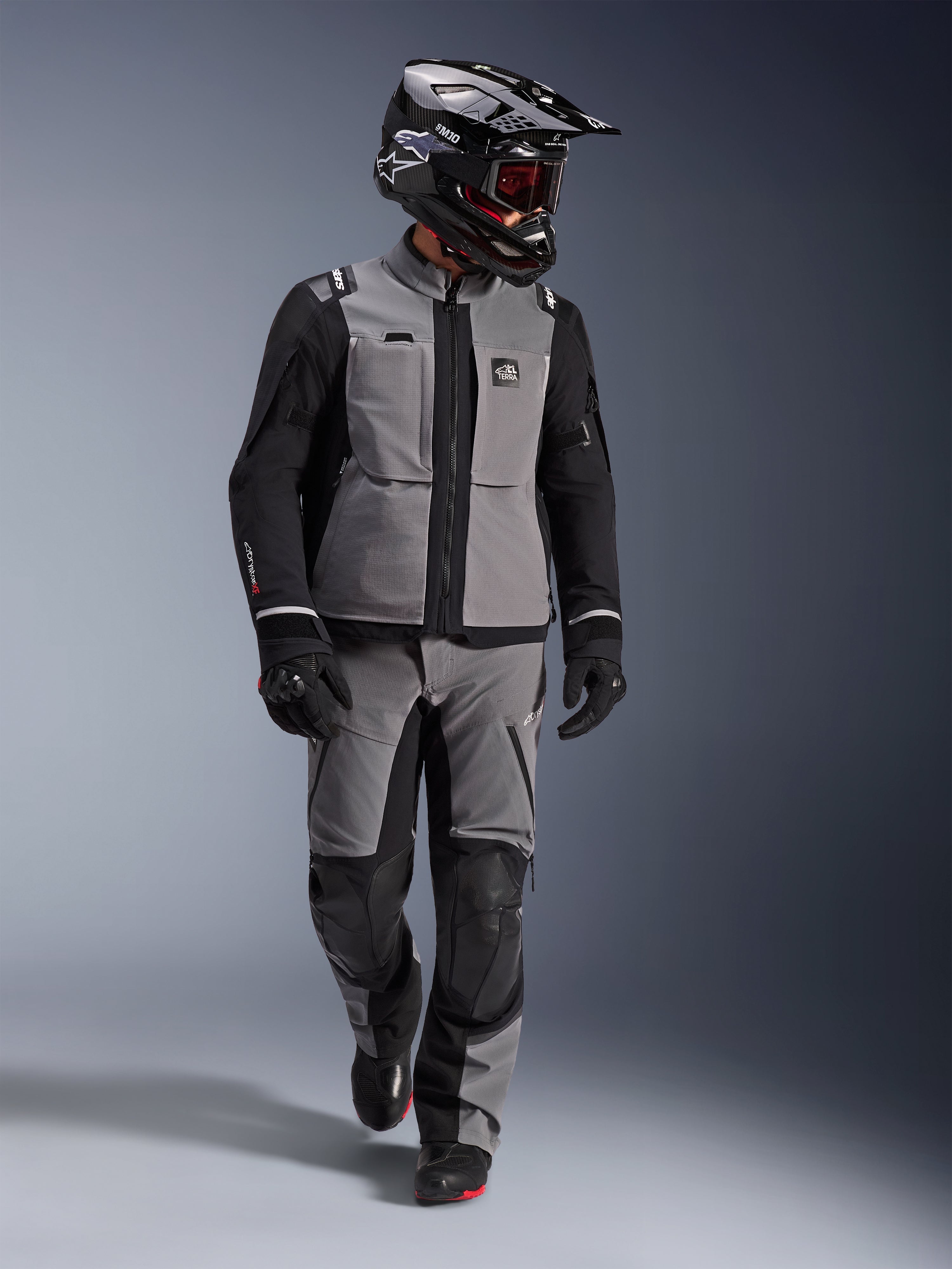 Motociclista de aventura con chaqueta Alpinestars Andes Pro Drystar® XF en gris oscuro y negro, pantalones de aventura a juego, casco Alpinestars negro con gafas, guantes y botas negros, equipamiento técnico completo de touring para viajes de larga distancia