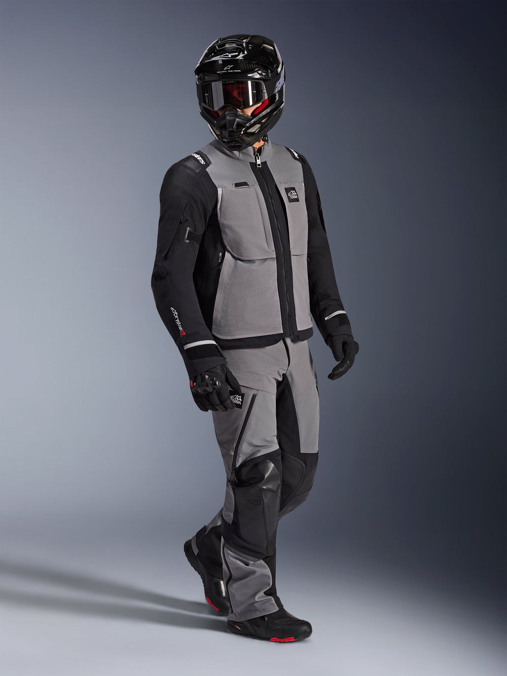 Motociclista de aventura con la chaqueta Alpinestars Andes Pro Drystar® XF en gris oscuro y negro, pantalones de aventura a juego en gris y negro, casco integral negro con gafas y guantes de moto negros, fotografía de estudio de equipamiento profesional para touring de larga distancia.