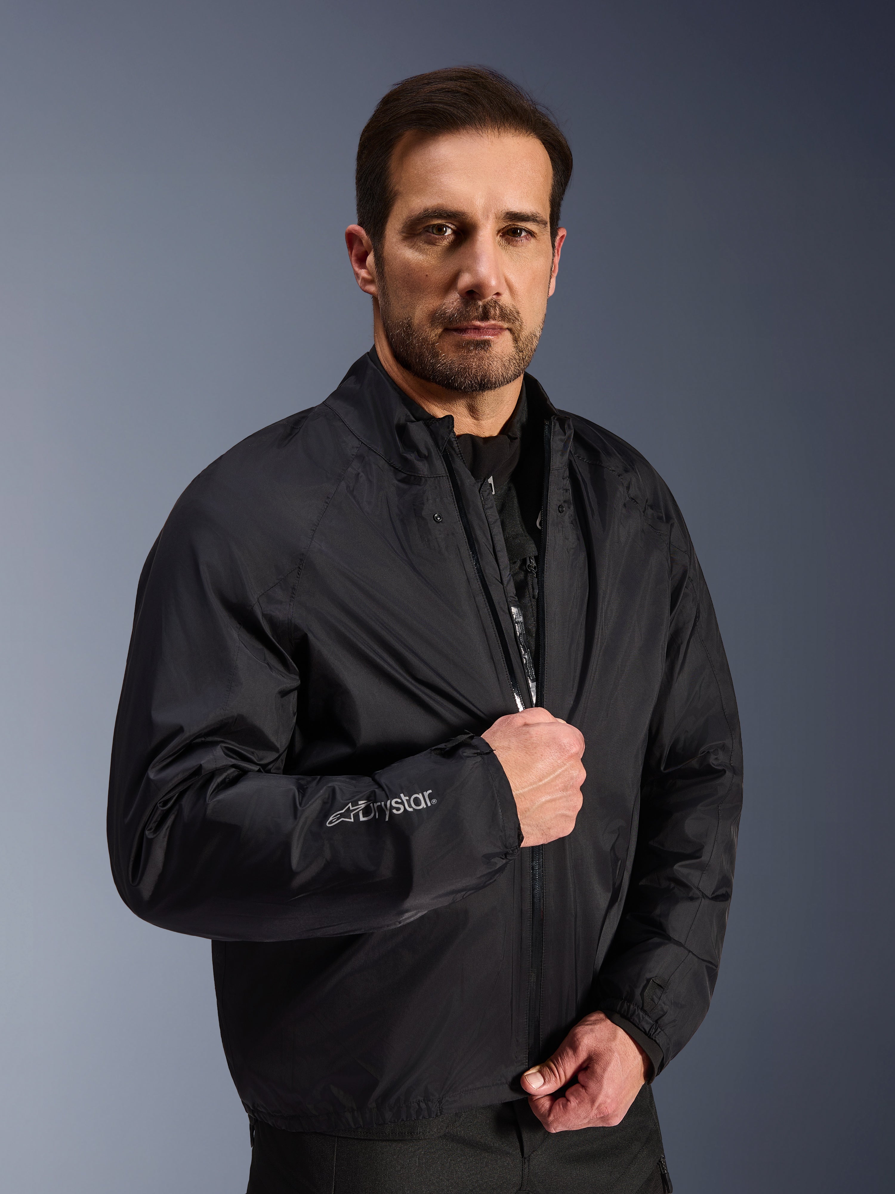 Chaqueta Alpinestars Cusco Drystar®, capa de membrana impermeable negra usada por un modelo masculino, con el logotipo Drystar en la manga, equipamiento ligero de motociclismo para adventure touring