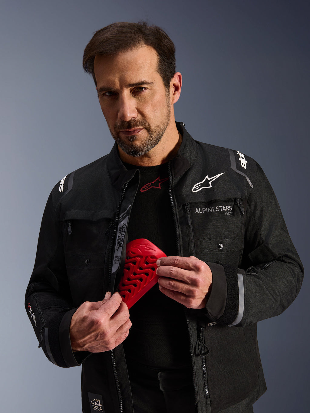 Chaqueta Alpinestars Cusco Drystar® en color negro, modelo masculino sosteniendo una pieza de protección Nucleon Flex Pro roja, chaqueta de moto para adventure touring con logotipos blancos, construcción de tejido técnico con cremalleras de ventilación y múltiples bolsillos