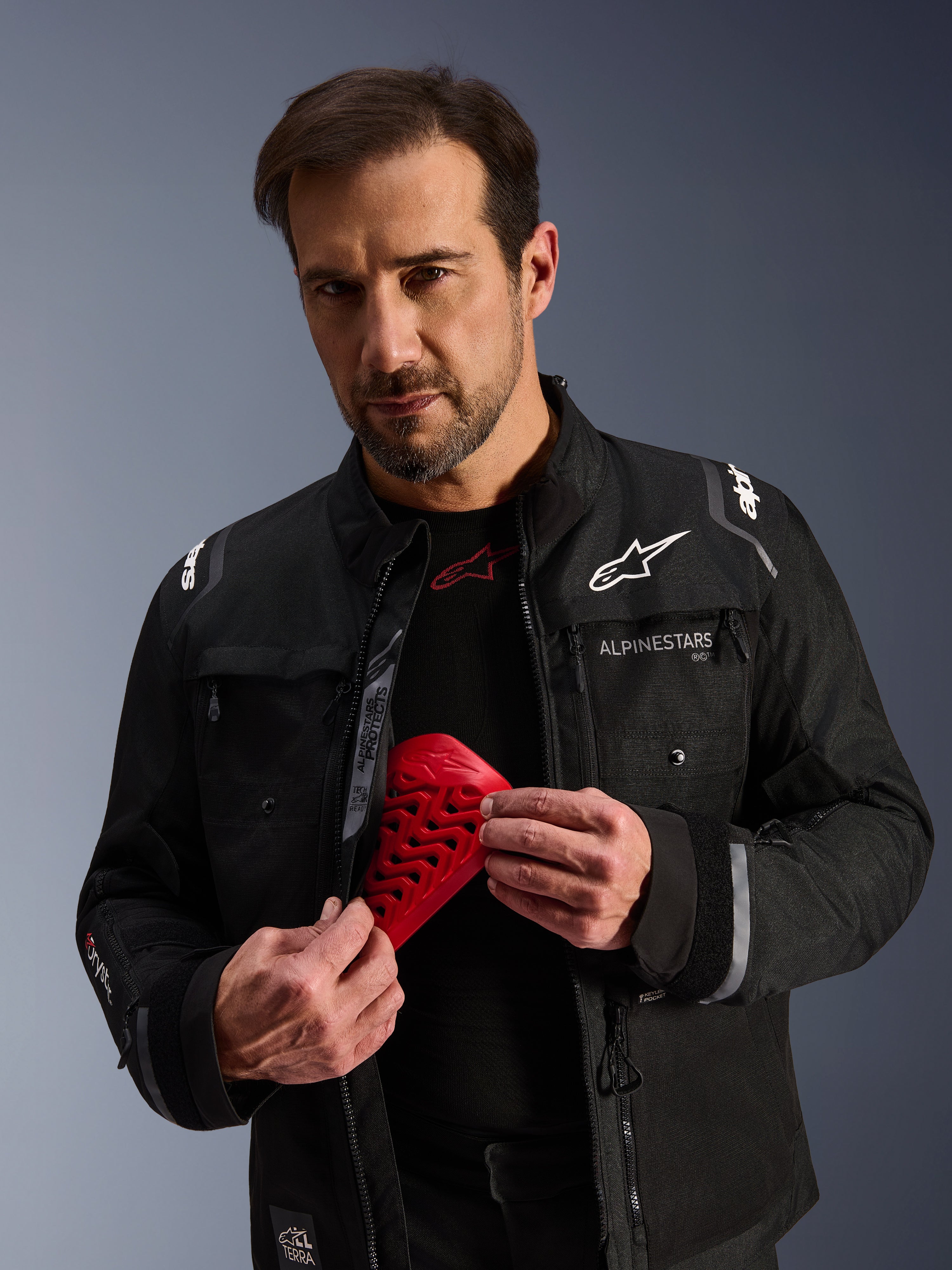 Chaqueta Alpinestars Cusco Drystar® en color negro, modelo masculino sosteniendo una pieza de protección Nucleon Flex Pro roja, chaqueta de moto para adventure touring con logotipos blancos, construcción de tejido técnico con cremalleras de ventilación y múltiples bolsillos