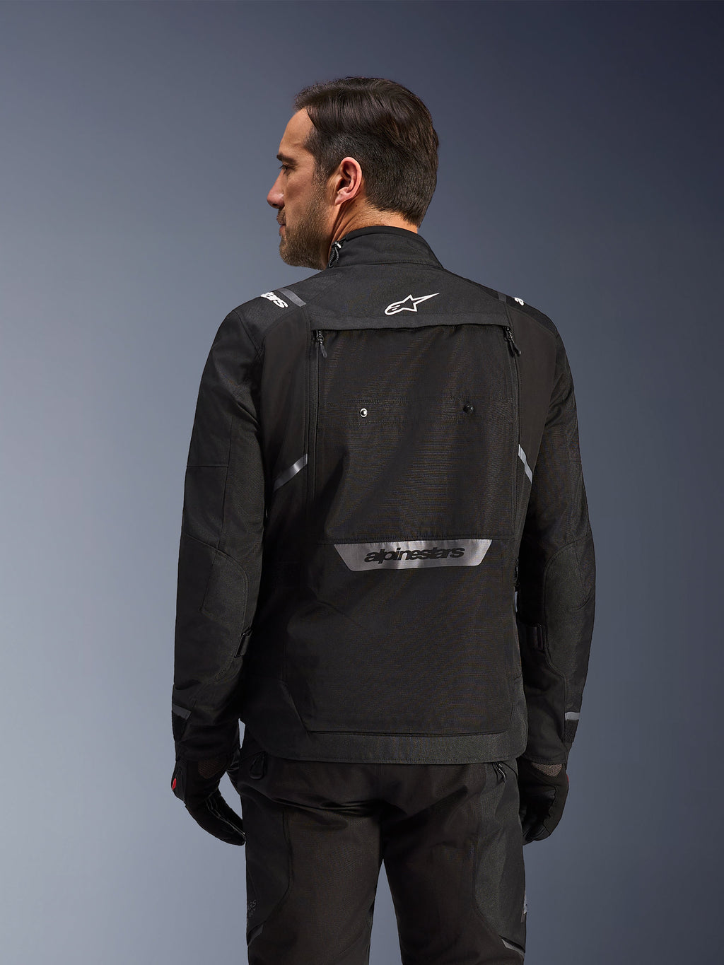 Motorista visto desde atrás con la chaqueta Alpinestars Cusco Drystar® en color negro, chaqueta textil de adventure touring con un gran bolsillo de almacenamiento trasero y logotipos reflectantes, combinada con pantalones y guantes negros a juego