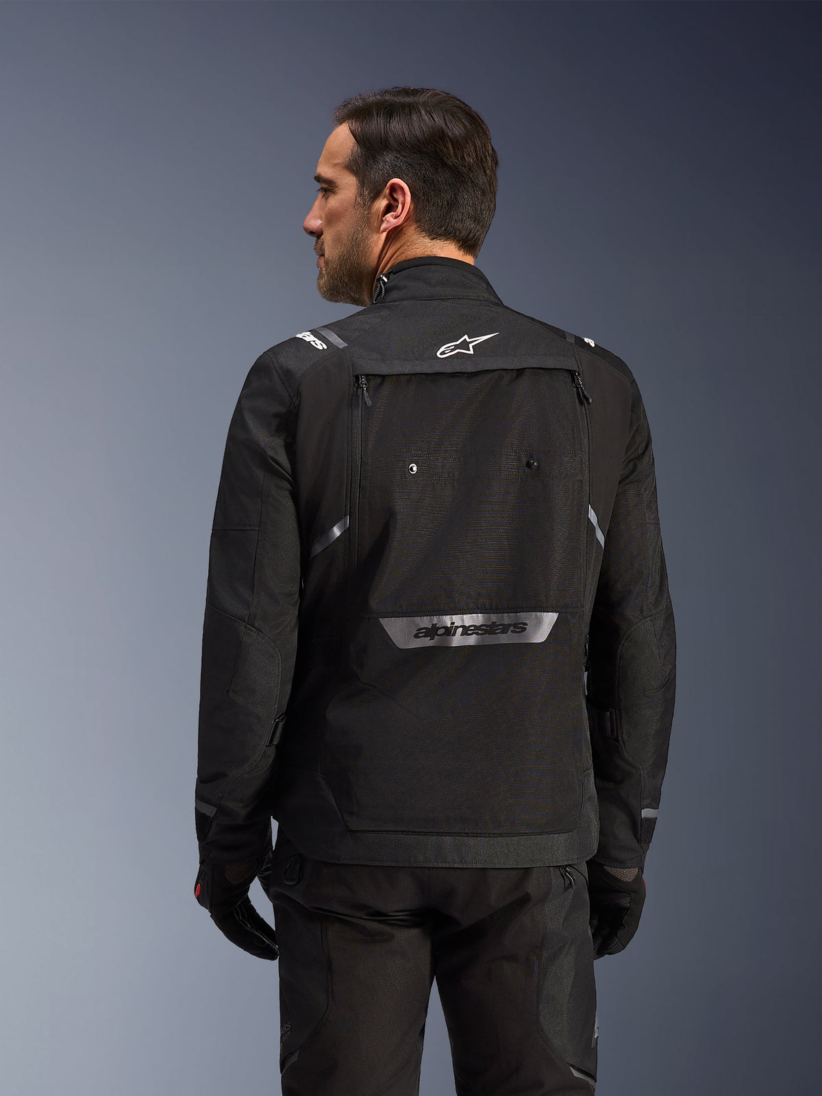 Motorista visto desde atrás con la chaqueta Alpinestars Cusco Drystar® en color negro, chaqueta textil de adventure touring con un gran bolsillo de almacenamiento trasero y logotipos reflectantes, combinada con pantalones y guantes negros a juego