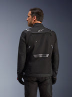 Motorista visto desde atrás con la chaqueta Alpinestars Cusco Drystar® en color negro, chaqueta textil de adventure touring con un gran bolsillo de almacenamiento trasero y logotipos reflectantes, combinada con pantalones y guantes negros a juego