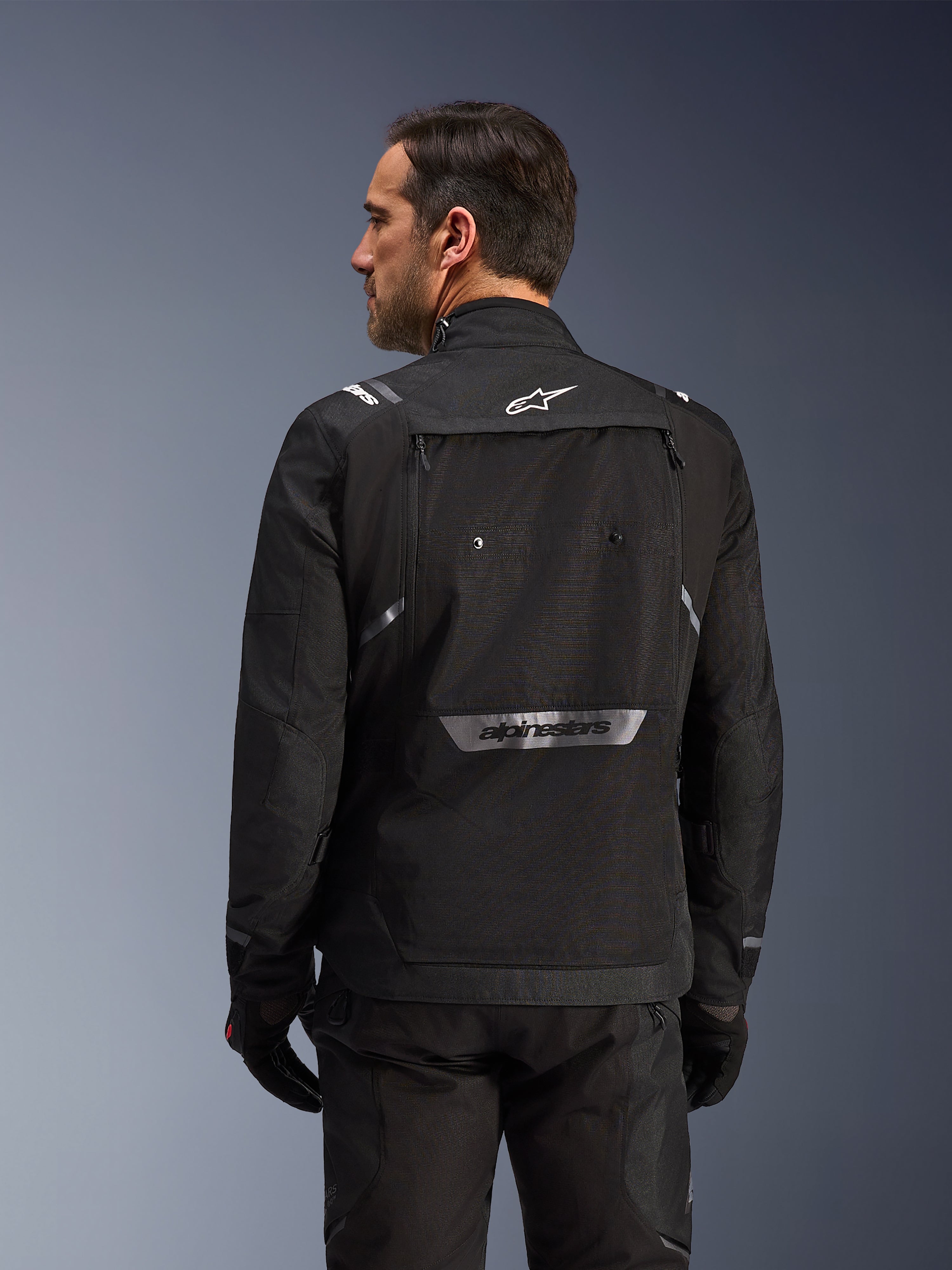 Motorista visto desde atrás con la chaqueta Alpinestars Cusco Drystar® en color negro, chaqueta textil de adventure touring con un gran bolsillo de almacenamiento trasero y logotipos reflectantes, combinada con pantalones y guantes negros a juego