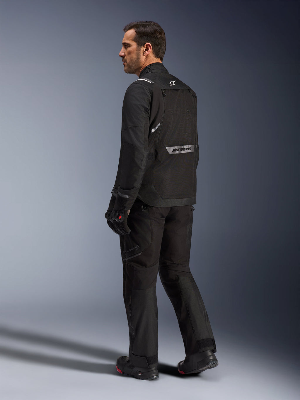 Chaqueta Alpinestars Cusco Drystar®, chaqueta de moto negra, vista posterior de un modelo masculino con equipamiento completo de conducción, con logotipos reflectantes en la espalda, construcción textil con paneles técnicos, conjunto coordinado para touring de aventura en carretera