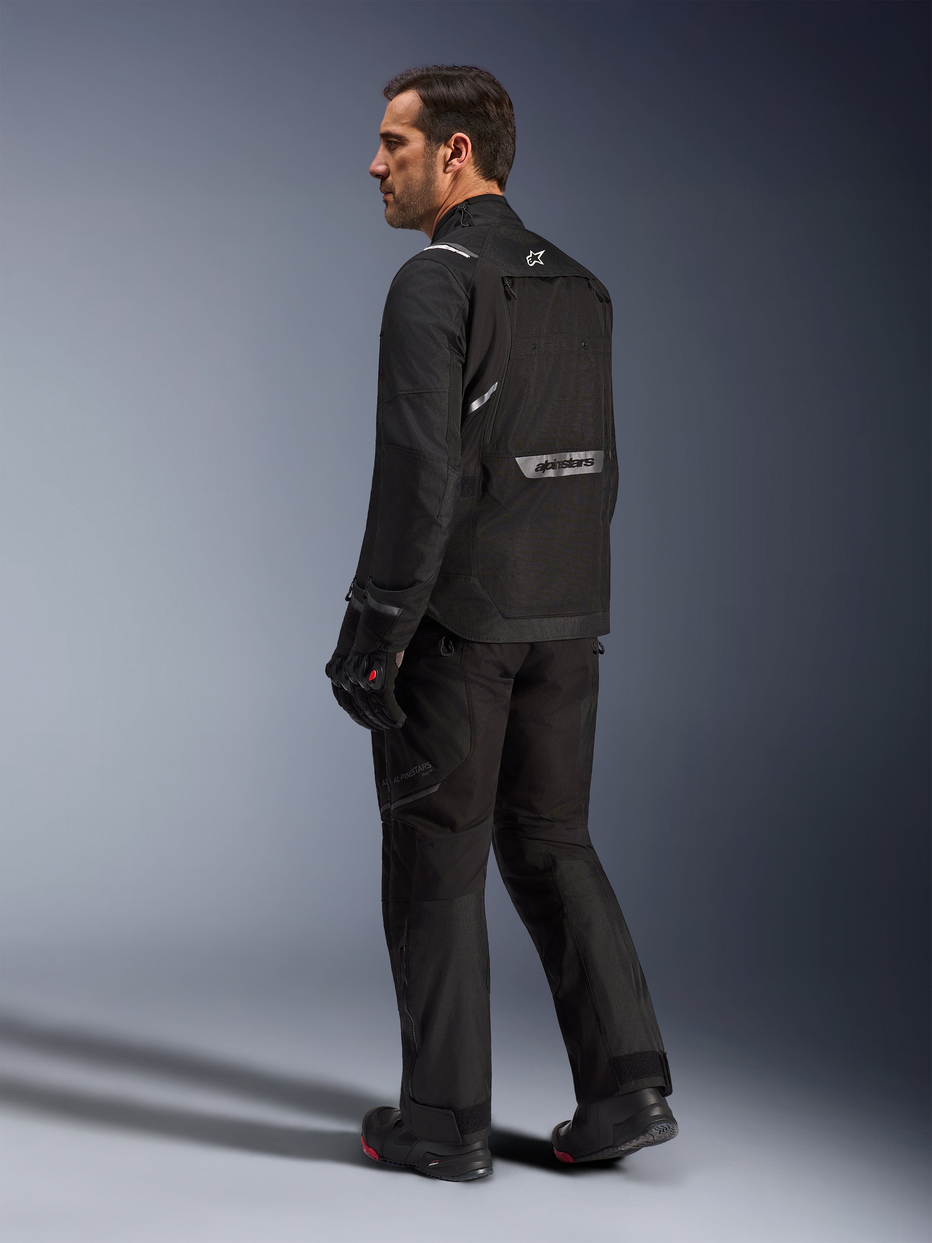 Chaqueta Alpinestars Cusco Drystar®, chaqueta de moto negra, vista posterior de un modelo masculino con equipamiento completo de conducción, con logotipos reflectantes en la espalda, construcción textil con paneles técnicos, conjunto coordinado para touring de aventura en carretera