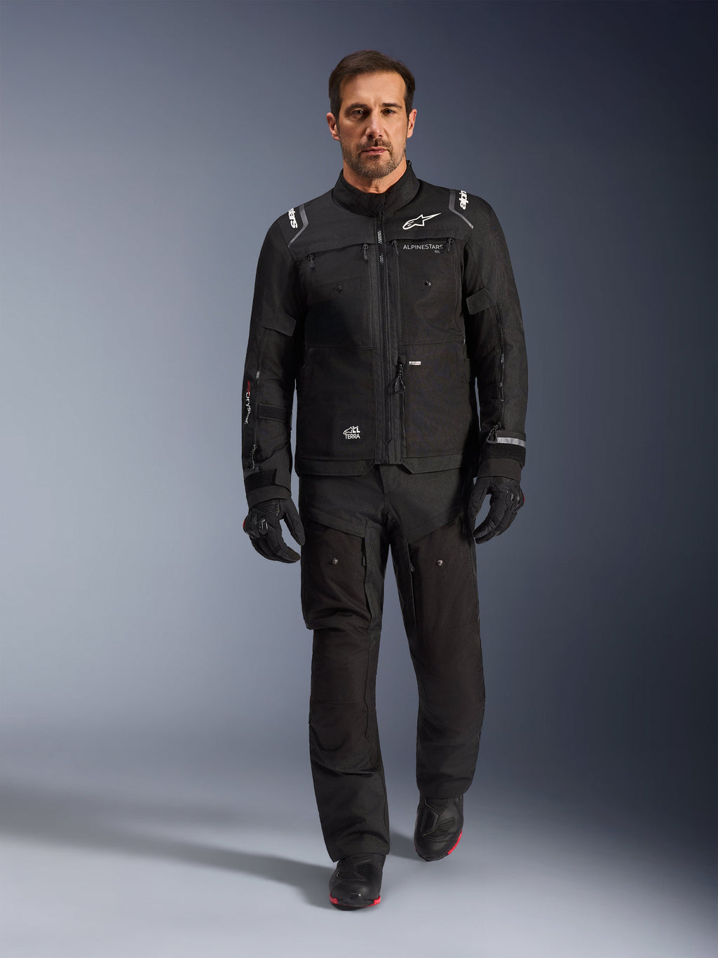 Piloto masculino vistiendo la chaqueta Alpinestars Cusco Drystar® en negro, combinada con pantalones técnicos de pilotaje y guantes negros a juego, toma de cuerpo completo en estudio, equipamiento de motociclismo adventure touring con marca Alpinestars visible y diseño multibolsillos