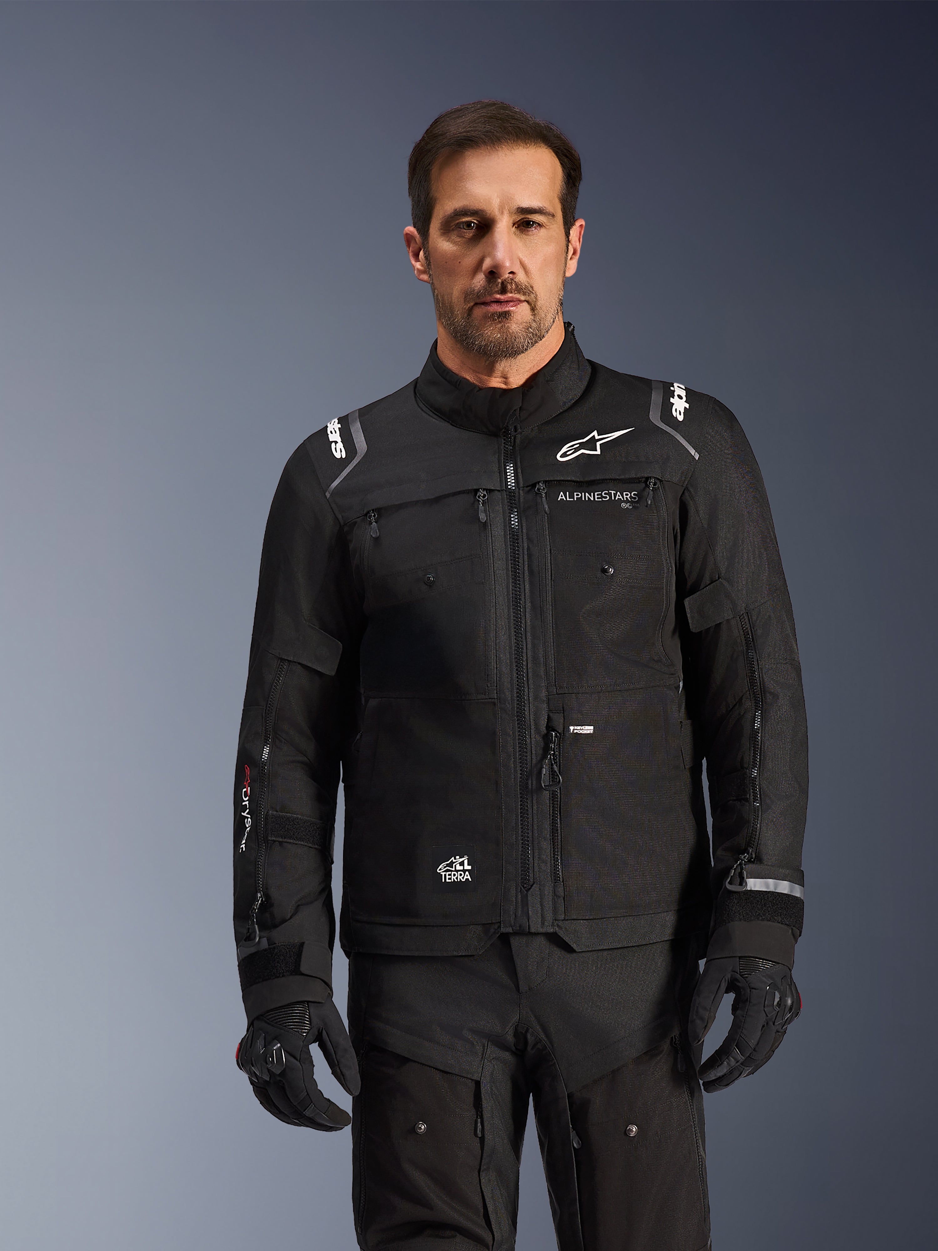 Chaqueta Cusco Drystar® de Alpinestars, chaqueta de motociclismo negra, vestida por un modelo con guantes y pantalones negros a juego, con construcción de tejido técnico y logotipos blancos de Alpinestars, diseñada para el adventure touring por carretera