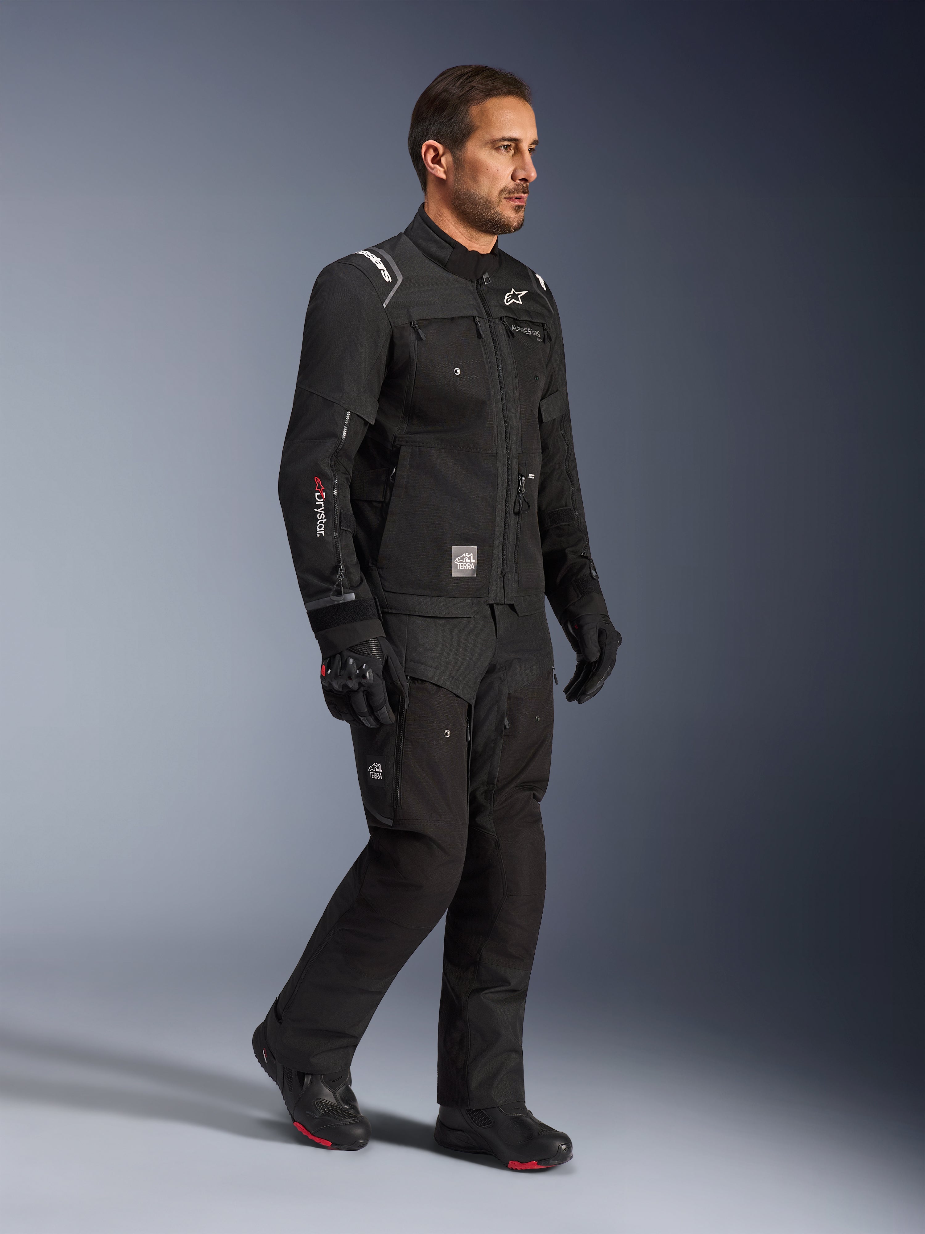 Modelo masculino vistiendo la chaqueta Alpinestars Cusco Drystar® en color negro, equipamiento de motociclismo adventure touring con pantalones y botas negras a juego, con un diseño multibolsillo, marca 'Drystar' en la manga y parche con el logo 'Terra'.