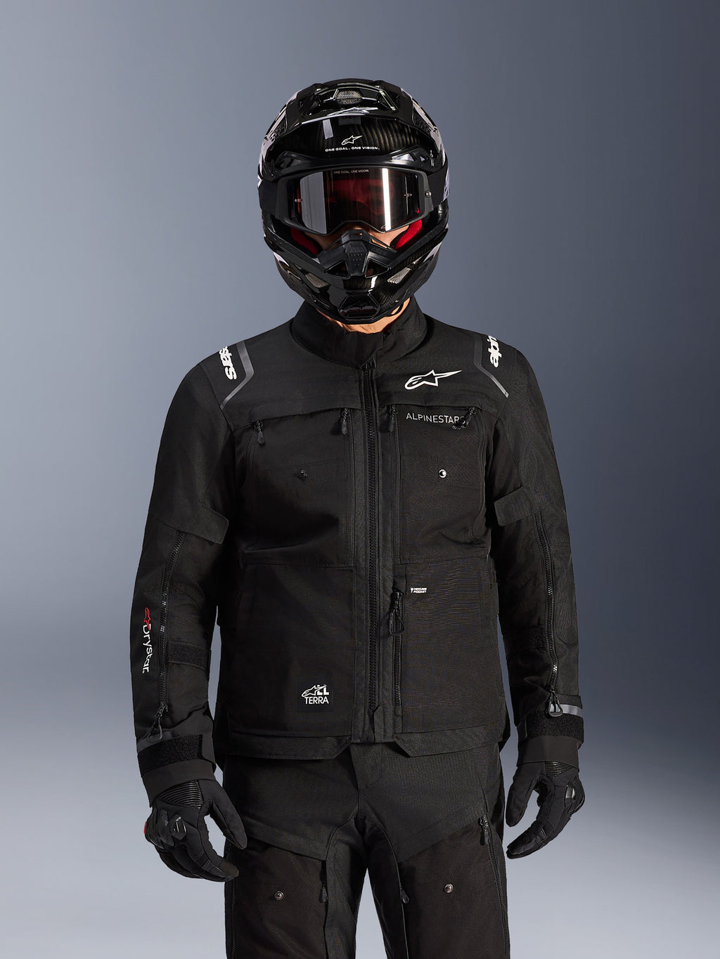 Chaqueta Alpinestars Cusco Drystar®, chaqueta negra de motociclismo adventure touring, combinada con un casco integral adventure negro y gafas oscuras, con múltiples bolsillos de utilidad, logotipo de Alpinestars en el pecho y logo Drystar® en la manga, parte de un equipamiento completo de aventura en carretera.