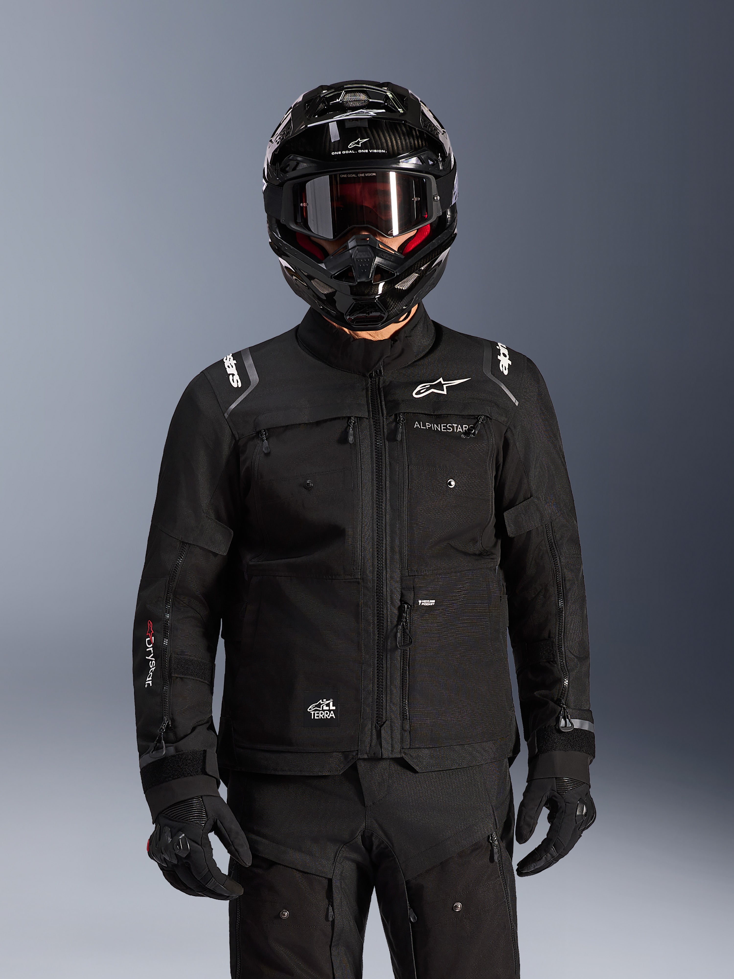 Chaqueta Alpinestars Cusco Drystar®, chaqueta negra de motociclismo adventure touring, combinada con un casco integral adventure negro y gafas oscuras, con múltiples bolsillos de utilidad, logotipo de Alpinestars en el pecho y logo Drystar® en la manga, parte de un equipamiento completo de aventura en carretera.