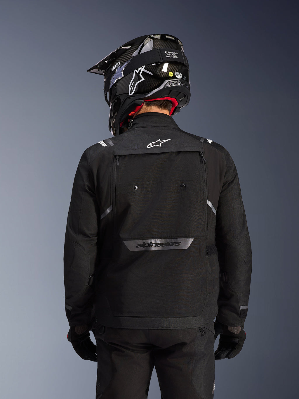 Chaqueta Alpinestars Cusco Drystar®, Negro, vista posterior de un piloto con casco y guantes negros, presenta amplios paneles de ventilación, logotipo blanco en el cuello y marca reflectante en la parte inferior de la espalda, equipamiento de adventure touring