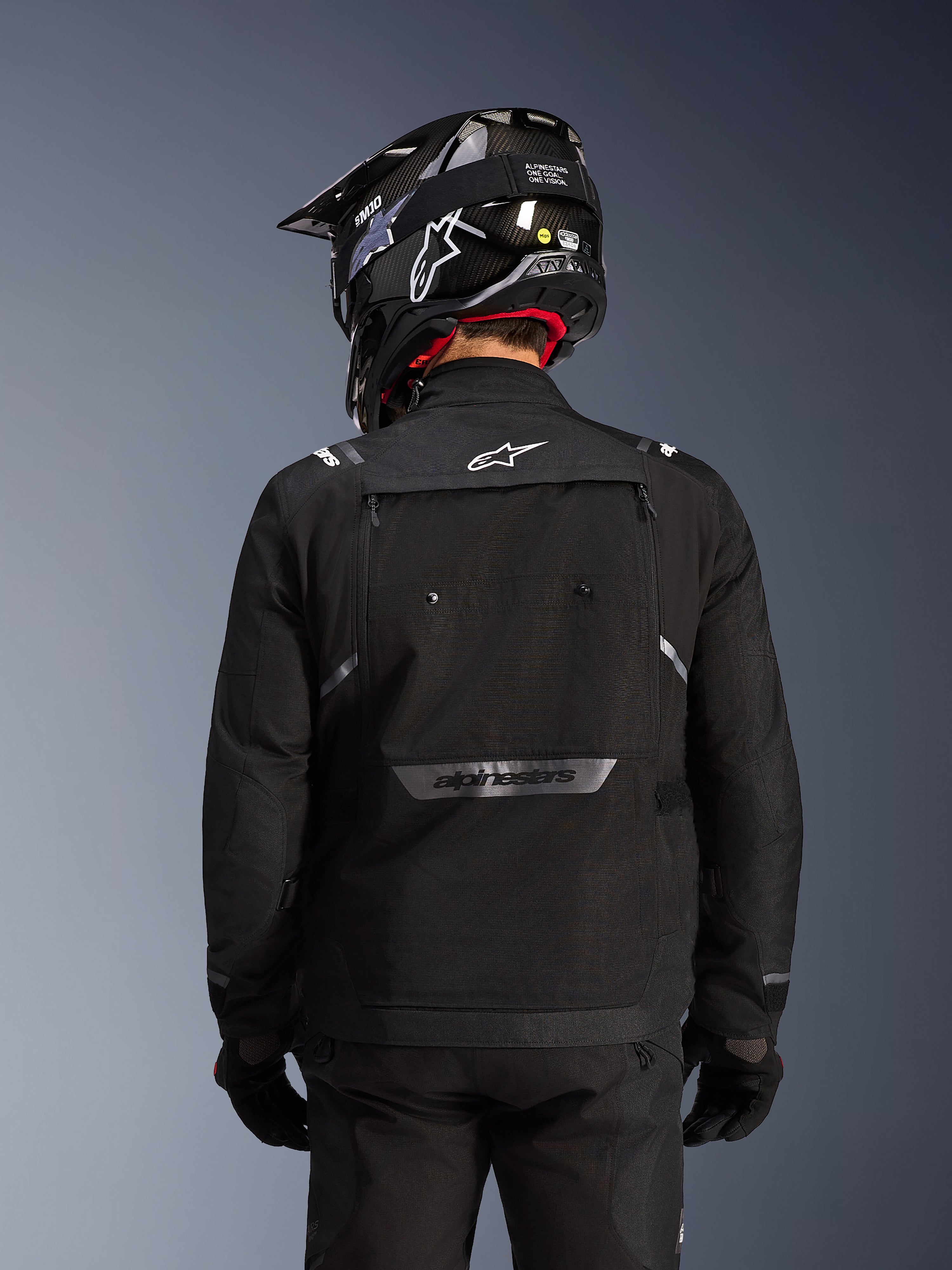 Chaqueta Alpinestars Cusco Drystar®, Negro, vista posterior de un piloto con casco y guantes negros, presenta amplios paneles de ventilación, logotipo blanco en el cuello y marca reflectante en la parte inferior de la espalda, equipamiento de adventure touring