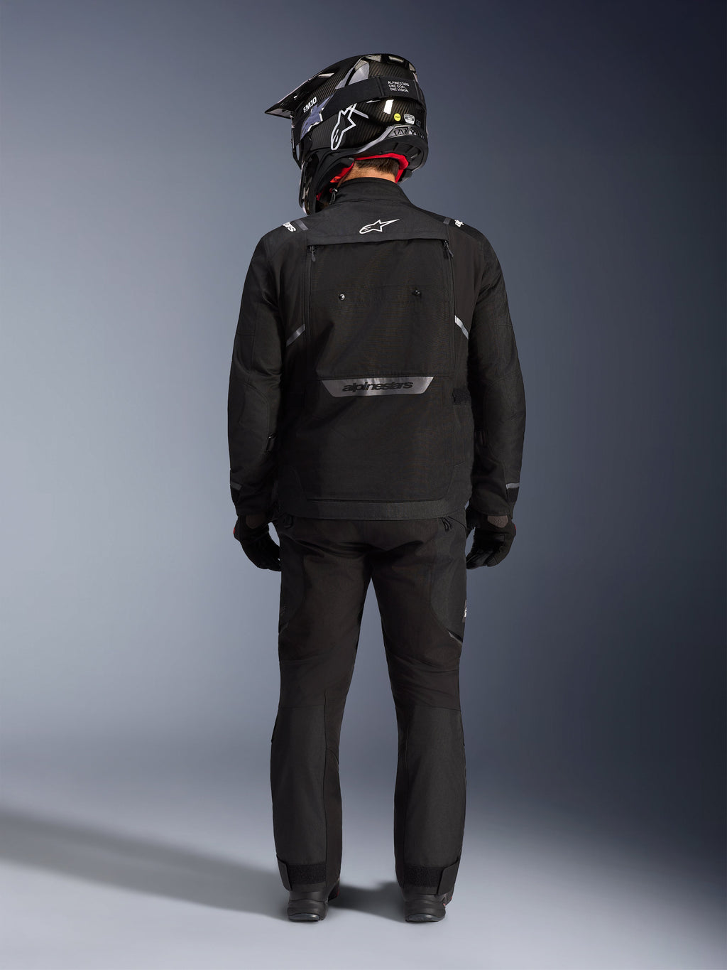 Chaqueta Alpinestars Cusco Drystar, chaqueta de aventura para motociclismo, negra, vista trasera combinada con pantalones a juego y un casco negro y blanco, con logotipos reflectantes de Alpinestars y grandes paneles de ventilación, construcción textil duradera para turismo de aventura por carretera