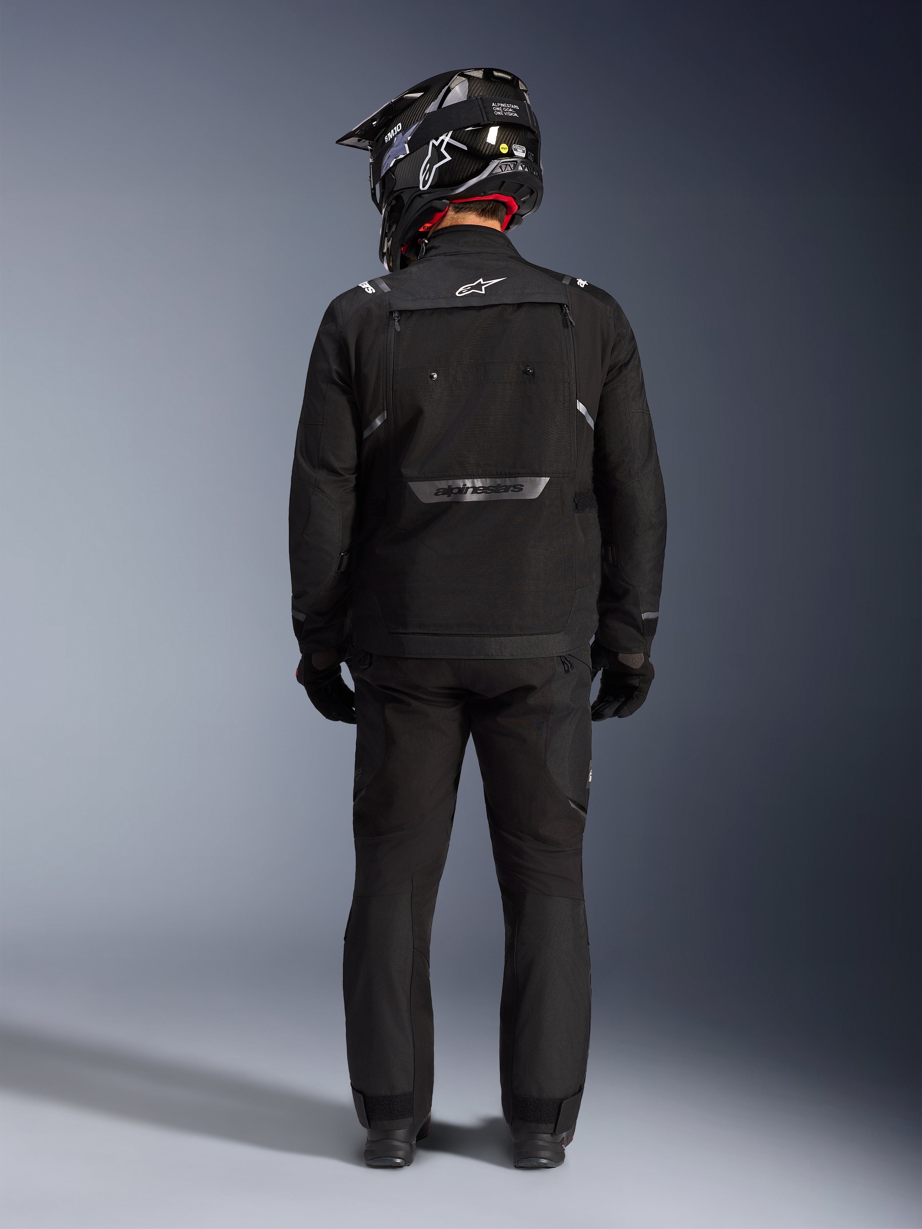 Chaqueta Alpinestars Cusco Drystar, chaqueta de aventura para motociclismo, negra, vista trasera combinada con pantalones a juego y un casco negro y blanco, con logotipos reflectantes de Alpinestars y grandes paneles de ventilación, construcción textil duradera para turismo de aventura por carretera