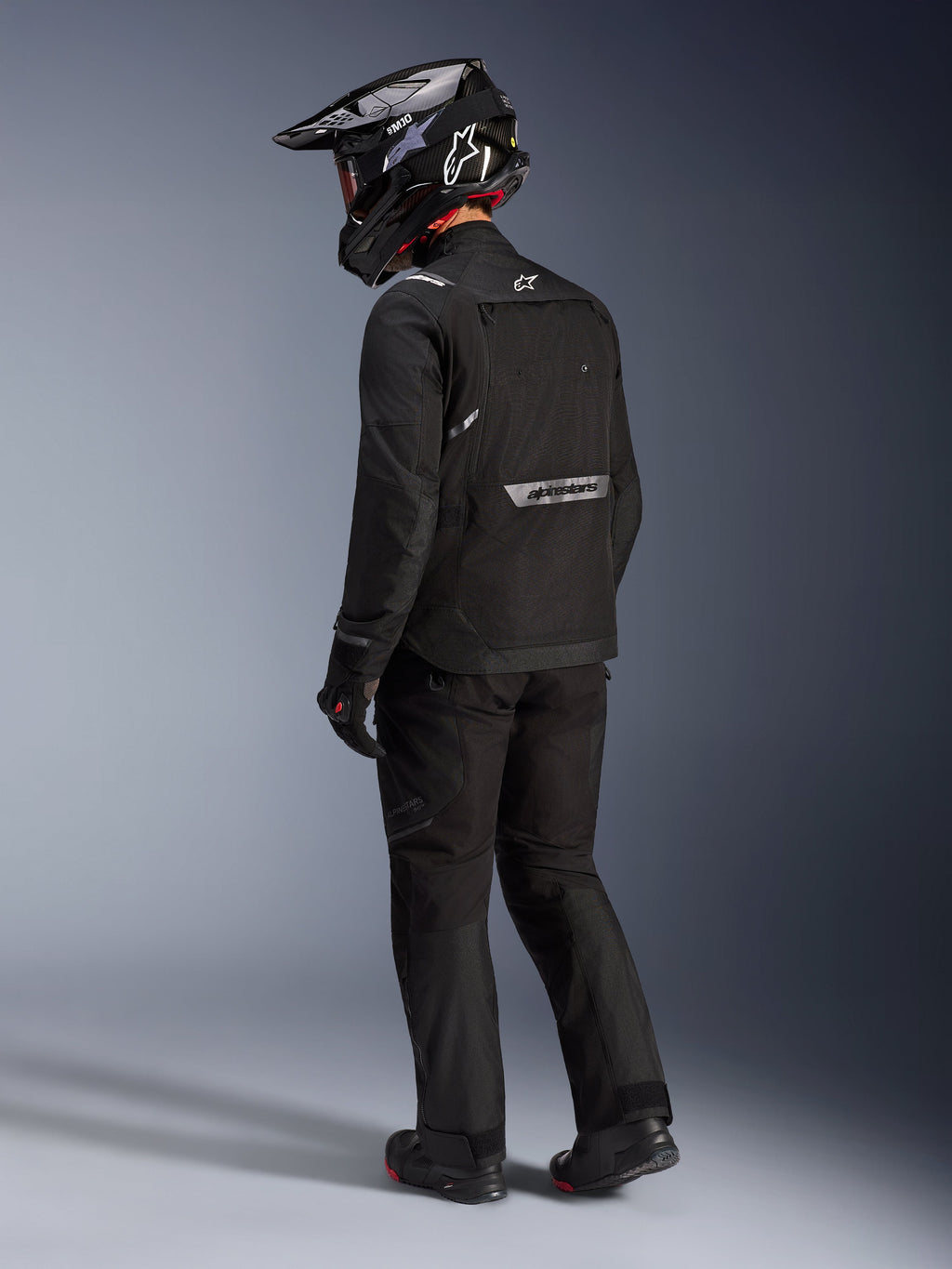 Chaqueta Alpinestars Cusco Drystar® en color negro, vista lateral trasera de un motorista con un conjunto completo de adventure touring que incluye pantalones y guantes negros a juego, casco de aventura negro, confección textil con logotipos reflectantes en la espalda