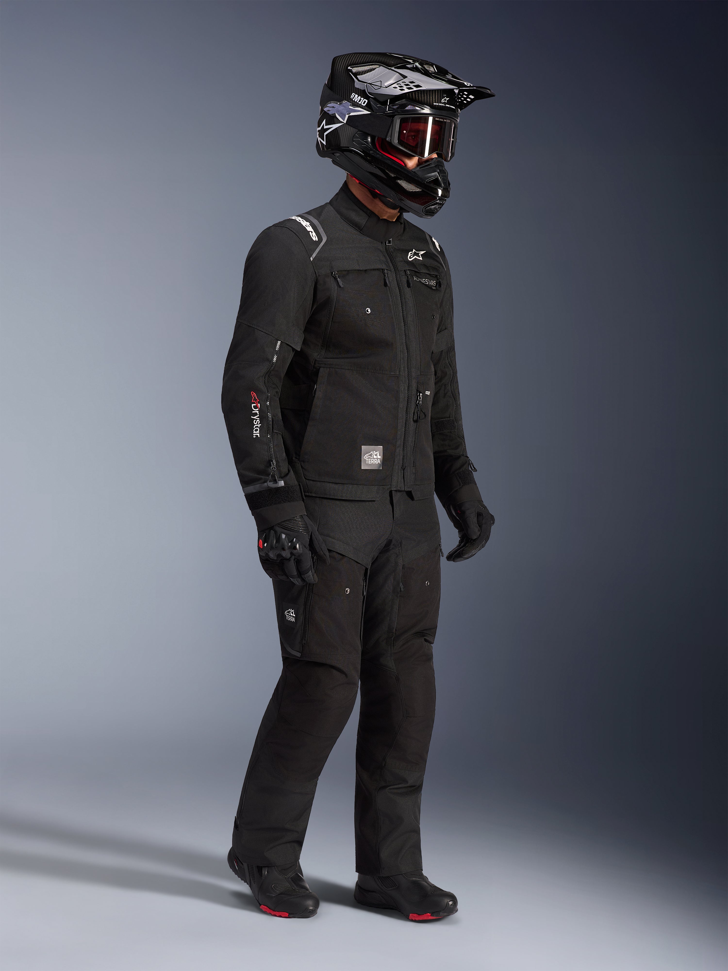 Chaqueta Alpinestars Cusco Drystar®, chaqueta de motociclismo adventure touring negra, usada con pantalones negros a juego y casco integral con gafas, con diseño funcional de múltiples bolsillos, construcción en tejido técnico y marca Drystar en la manga.