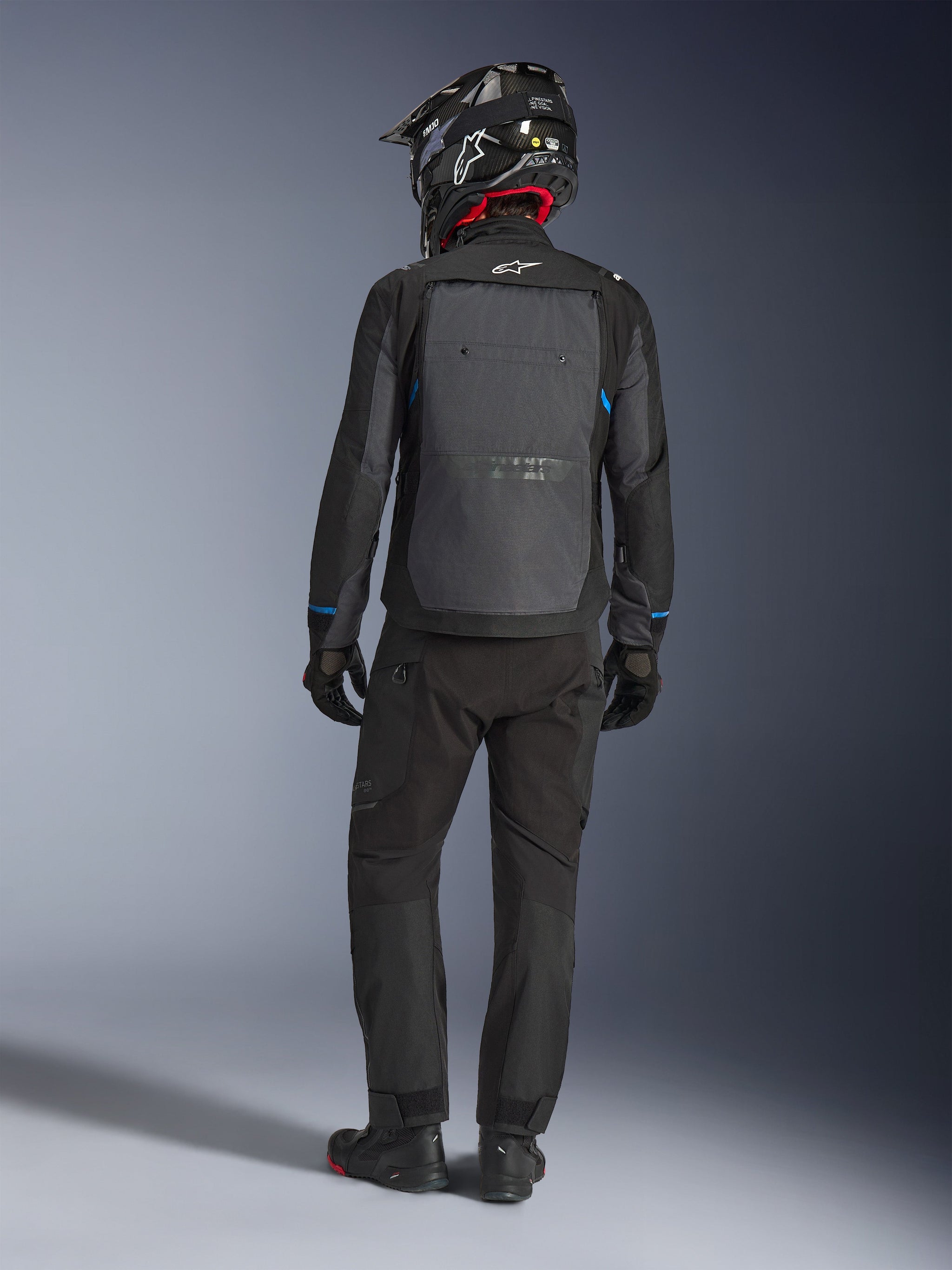 Chaqueta Alpinestars Cusco Drystar®, negro y gris oscuro, vista trasera de un piloto con equipamiento completo de adventure touring, gran panel trasero gris con el logotipo de Alpinestars, detalles azules en las mangas, usada con pantalones negros y casco a juego.