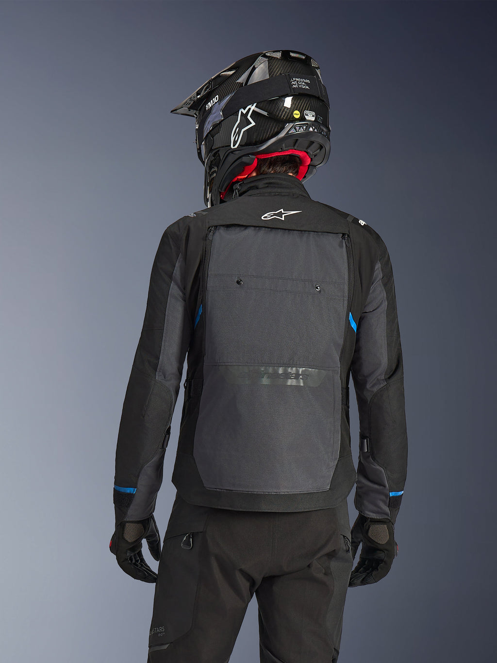 Motociclista visto desde atrás con la chaqueta Alpinestars Cusco Drystar® en color negro y gris oscuro, con un gran panel de ventilación trasero gris y logo blanco, combinado con un casco off-road negro y equipamiento de conducción para adventure touring.