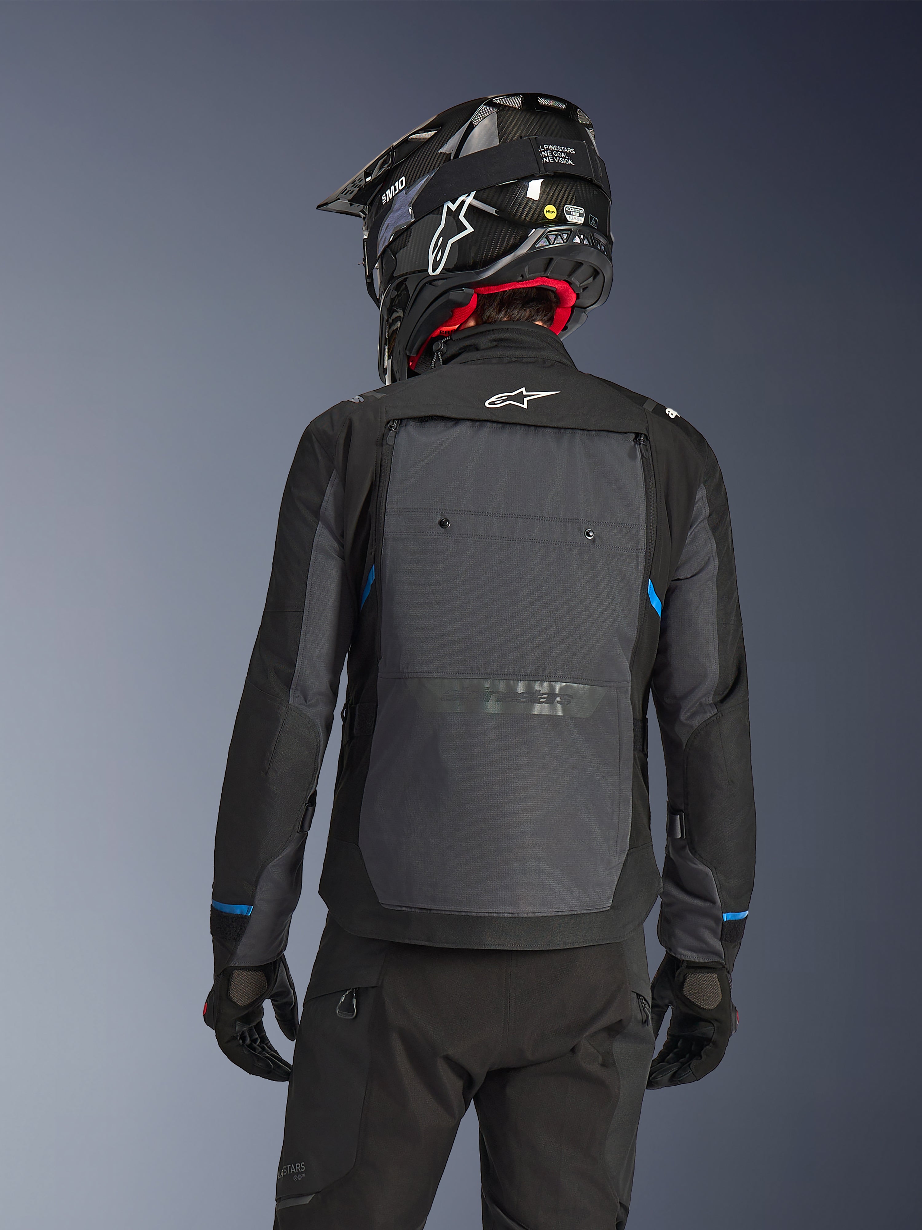 Motociclista visto desde atrás con la chaqueta Alpinestars Cusco Drystar® en color negro y gris oscuro, con un gran panel de ventilación trasero gris y logo blanco, combinado con un casco off-road negro y equipamiento de conducción para adventure touring.