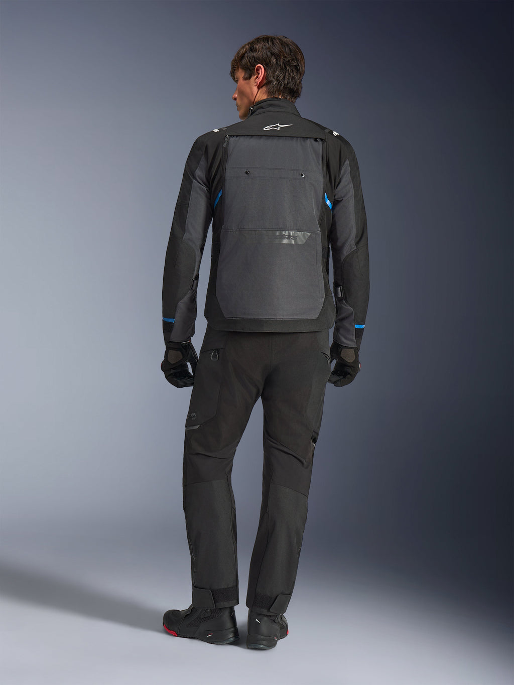 Chaqueta Alpinestars Cusco Drystar®, negro y gris oscuro con detalles azules, vista posterior, usada con pantalones de aventura y guantes negros a juego, cuenta con un gran panel gris en la espalda y el logotipo blanco en el cuello, diseñada para road y adventure touring