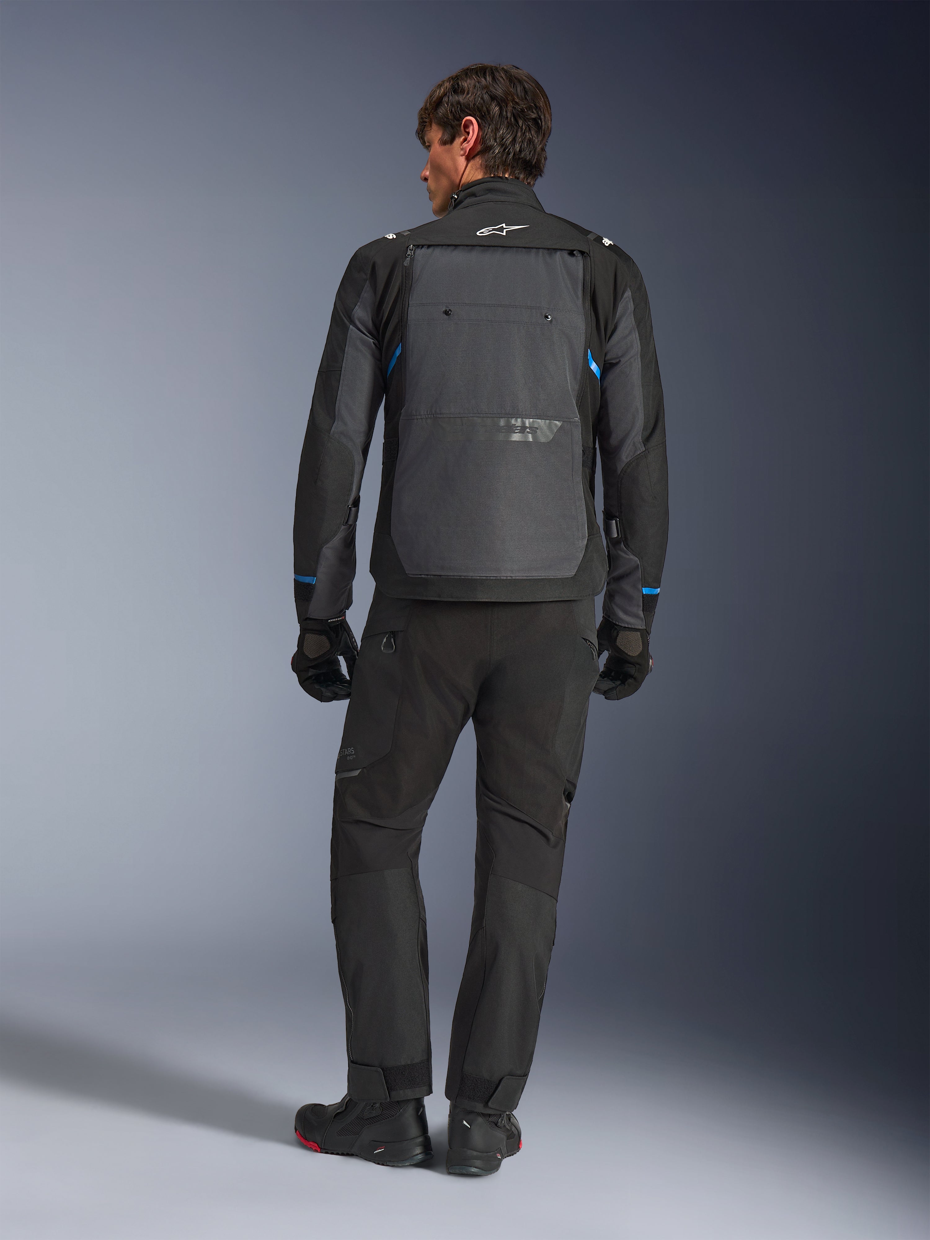 Chaqueta Alpinestars Cusco Drystar®, negro y gris oscuro con detalles azules, vista posterior, usada con pantalones de aventura y guantes negros a juego, cuenta con un gran panel gris en la espalda y el logotipo blanco en el cuello, diseñada para road y adventure touring