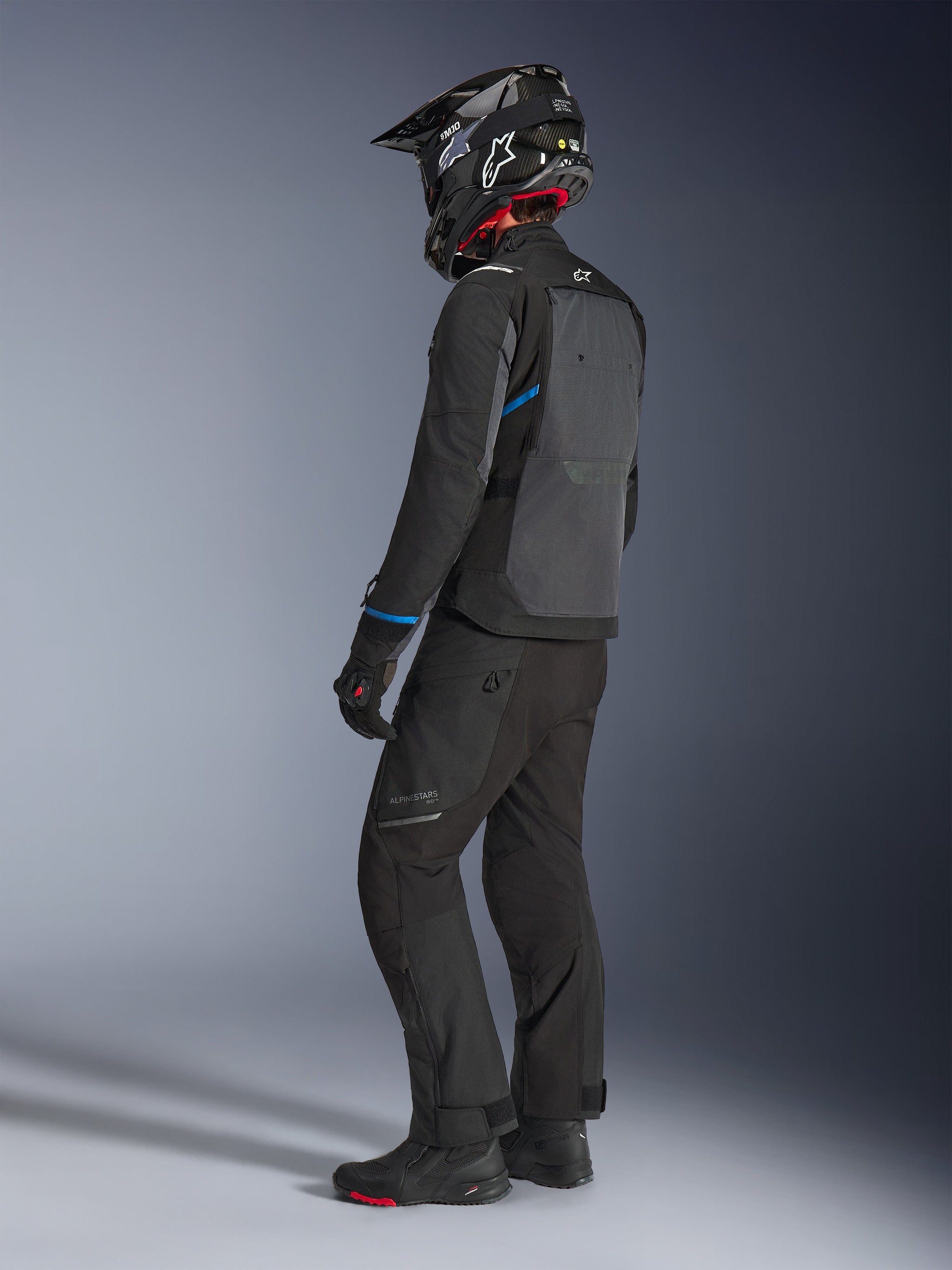 Motociclista vistiendo la chaqueta Alpinestars Cusco Drystar® en negro y gris oscuro con detalles azules, combinada con pantalones de motociclismo negros, casco SM10 de fibra de carbono y guantes negros, vista posterior que muestra el gran panel de ventilación y el diseño adventure touring.