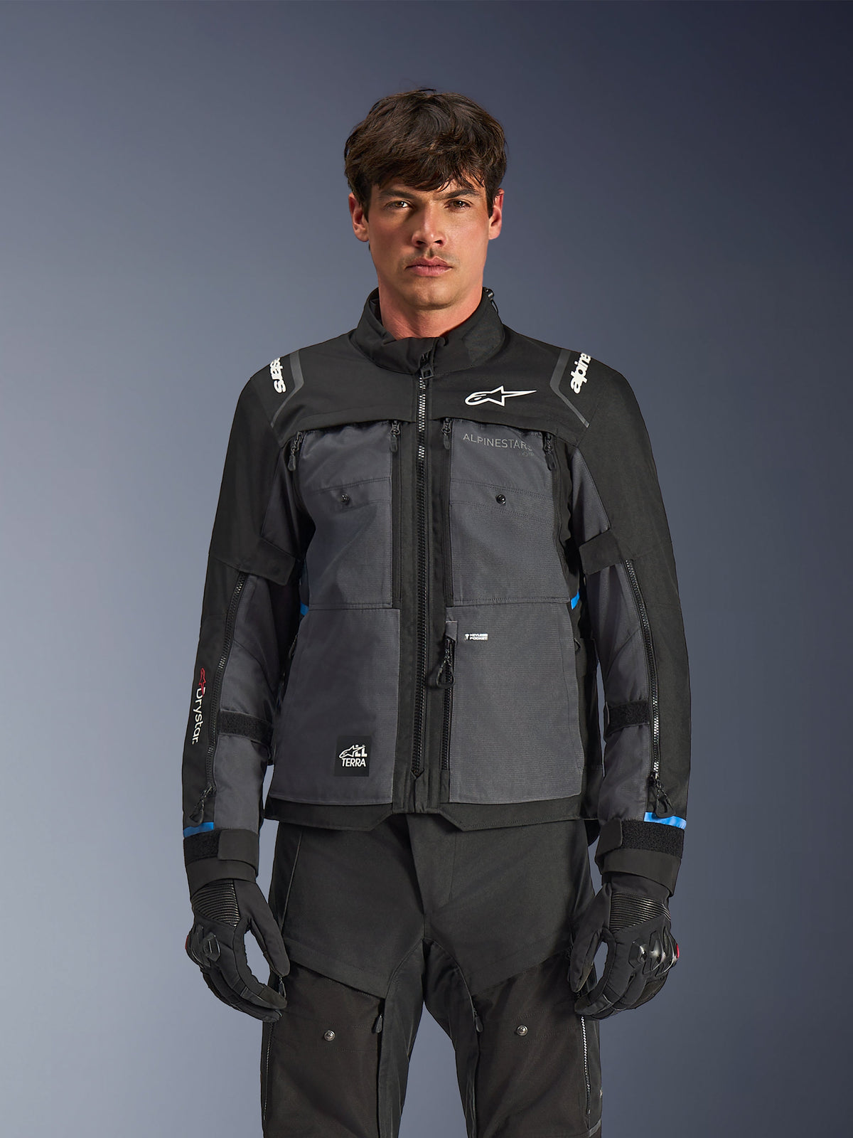 Chaqueta Alpinestars Cusco Drystar®, chaqueta de mototurismo, negro y gris oscuro, vista frontal en modelo, con logotipos de Alpinestars en el pecho y los hombros, marca Drystar® en la manga, diseño técnico multibolsillo con cremalleras de ventilación, combinada con equipamiento de conducción negro