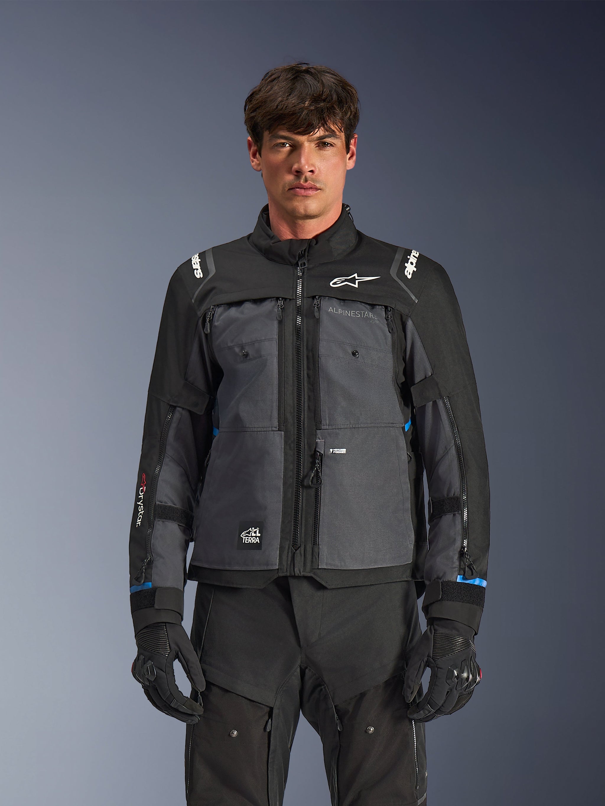Chaqueta Alpinestars Cusco Drystar®, chaqueta de mototurismo, negro y gris oscuro, vista frontal en modelo, con logotipos de Alpinestars en el pecho y los hombros, marca Drystar® en la manga, diseño técnico multibolsillo con cremalleras de ventilación, combinada con equipamiento de conducción negro