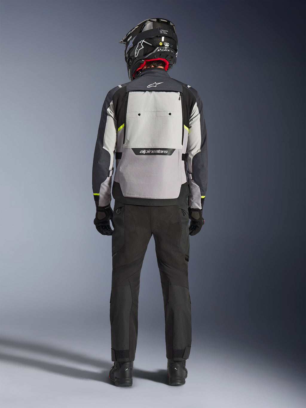 Motociclista visto desde atrás vistiendo la chaqueta Alpinestars Cusco Drystar® en Ice Gray, Mid Gray y Deep Gray, con un gran panel de ventilación trasero y marca Alpinestars, combinada con pantalones de moto negros y un casco negro, ropa de motociclismo para adventure touring.