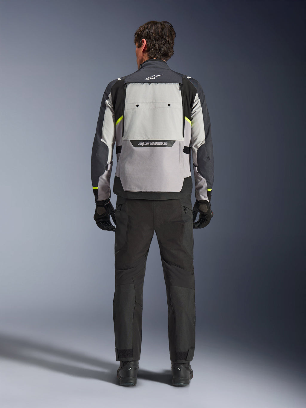 Vista trasera de un motociclista con la chaqueta Alpinestars Cusco Drystar® en color Ice Gray, Mid Gray y Deep Gray, combinada con pantalones y guantes de motociclismo negros, destacando el gran panel de ventilación trasero y el logotipo de Alpinestars.