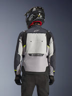 Chaqueta Alpinestars Cusco Drystar®, vista trasera, colores Ice Gray, Mid Gray y Deep Gray, con un gran panel de ventilación trasero y detalles amarillos de alta visibilidad, combinada con casco adventure negro y pantalones negros.