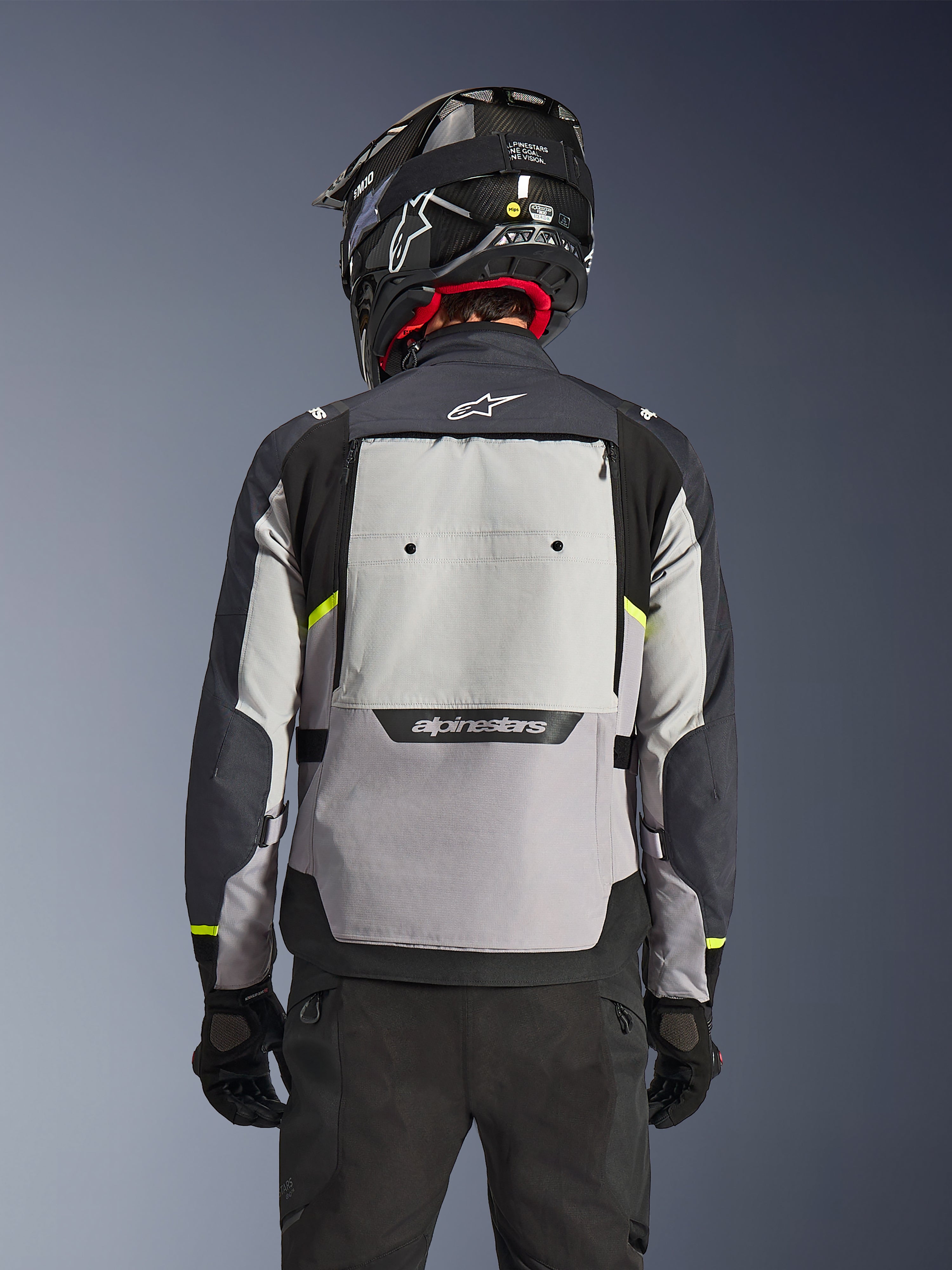 Chaqueta Alpinestars Cusco Drystar®, vista trasera, colores Ice Gray, Mid Gray y Deep Gray, con un gran panel de ventilación trasero y detalles amarillos de alta visibilidad, combinada con casco adventure negro y pantalones negros.