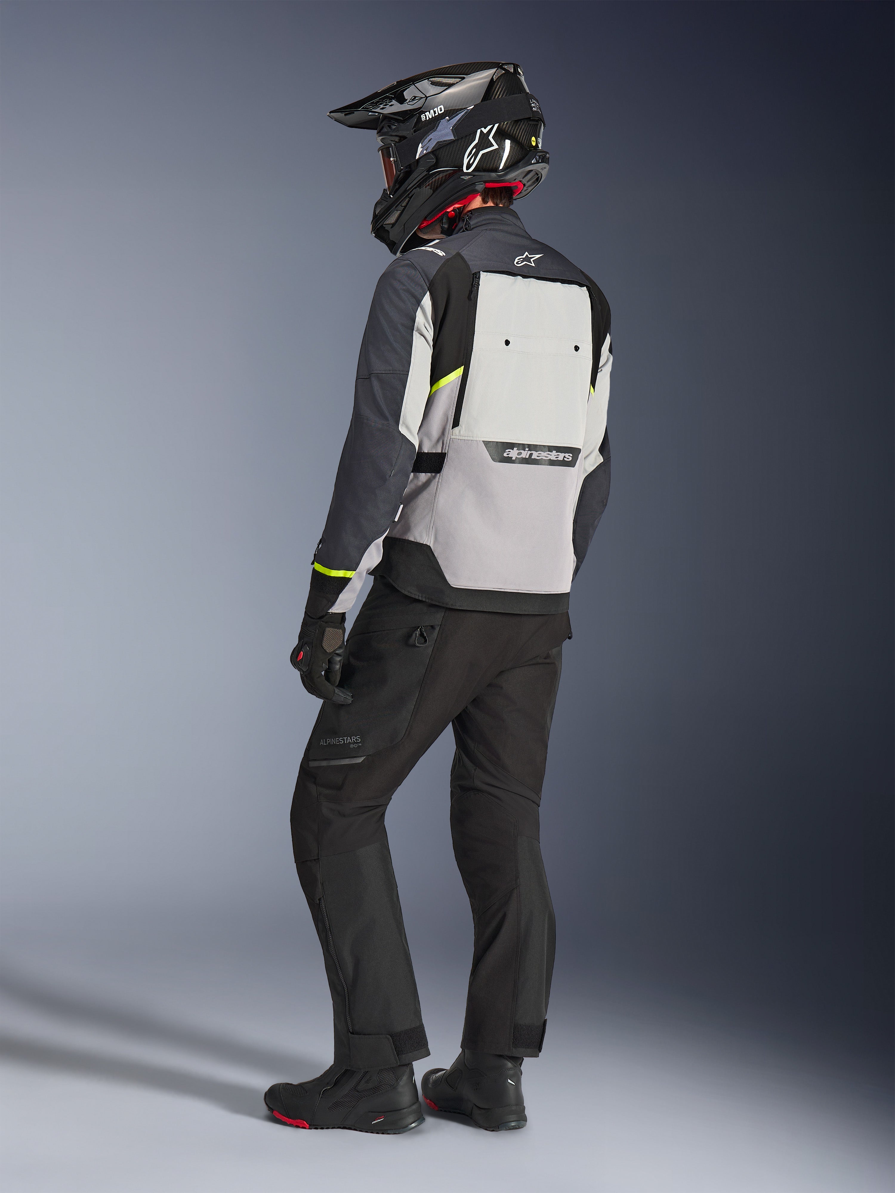 Chaqueta Alpinestars Cusco Drystar® en gris hielo, gris medio y gris oscuro, vista posterior en un motociclista. Presenta detalles en amarillo fluorescente, un amplio panel de ventilación trasero con la marca y tejido resistente. Se complementa con pantalones de moto negros y un casco de aventura para un look de touring completo.