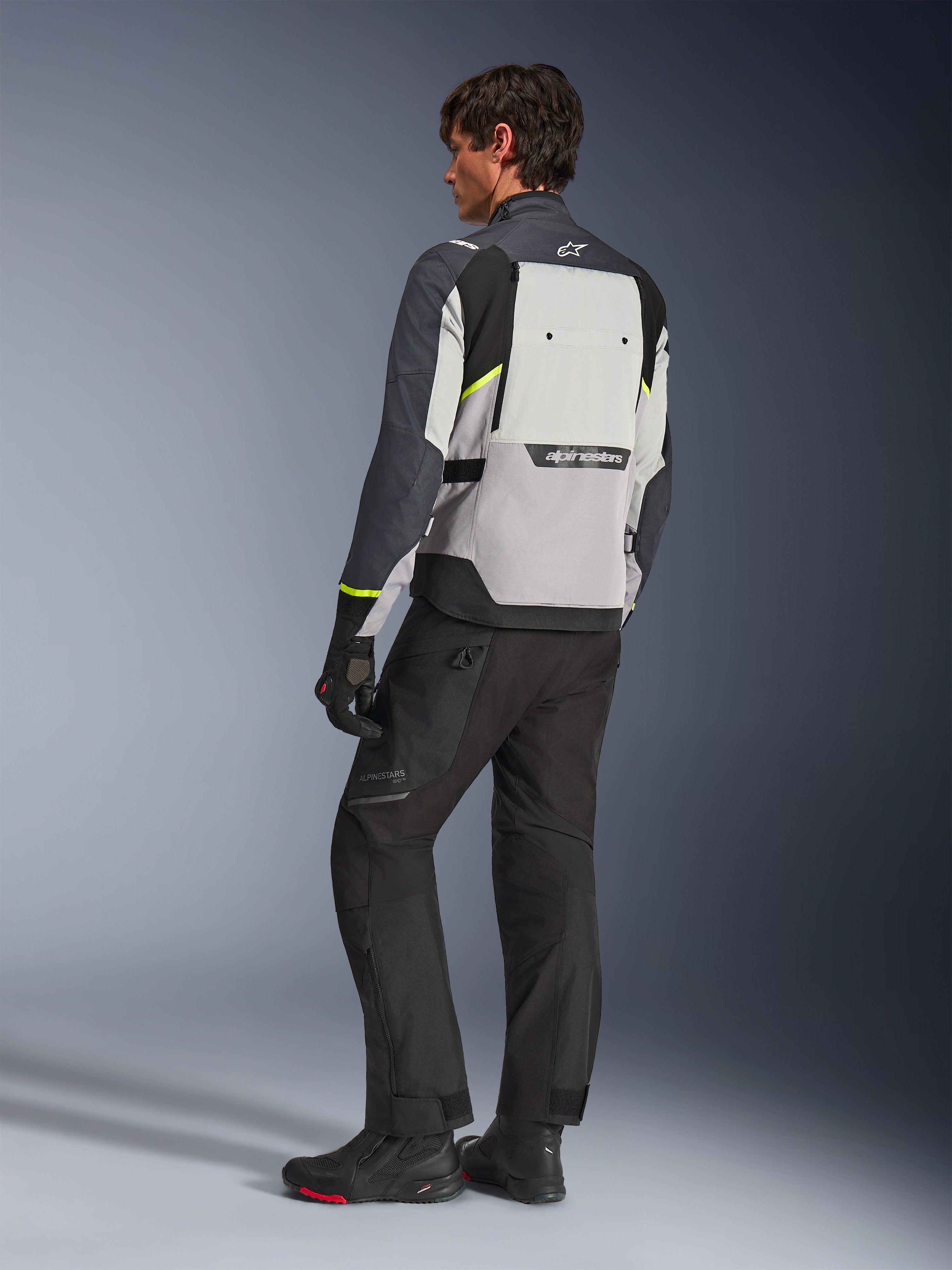 Chaqueta Alpinestars Cusco Drystar® en Gris Hielo, Gris Medio y Gris Profundo, vista trasera en modelo, chaqueta adventure touring con detalles en amarillo neón, usada con pantalones y botas de motociclismo negros, con un gran panel de ventilación en la espalda y marca Alpinestars.