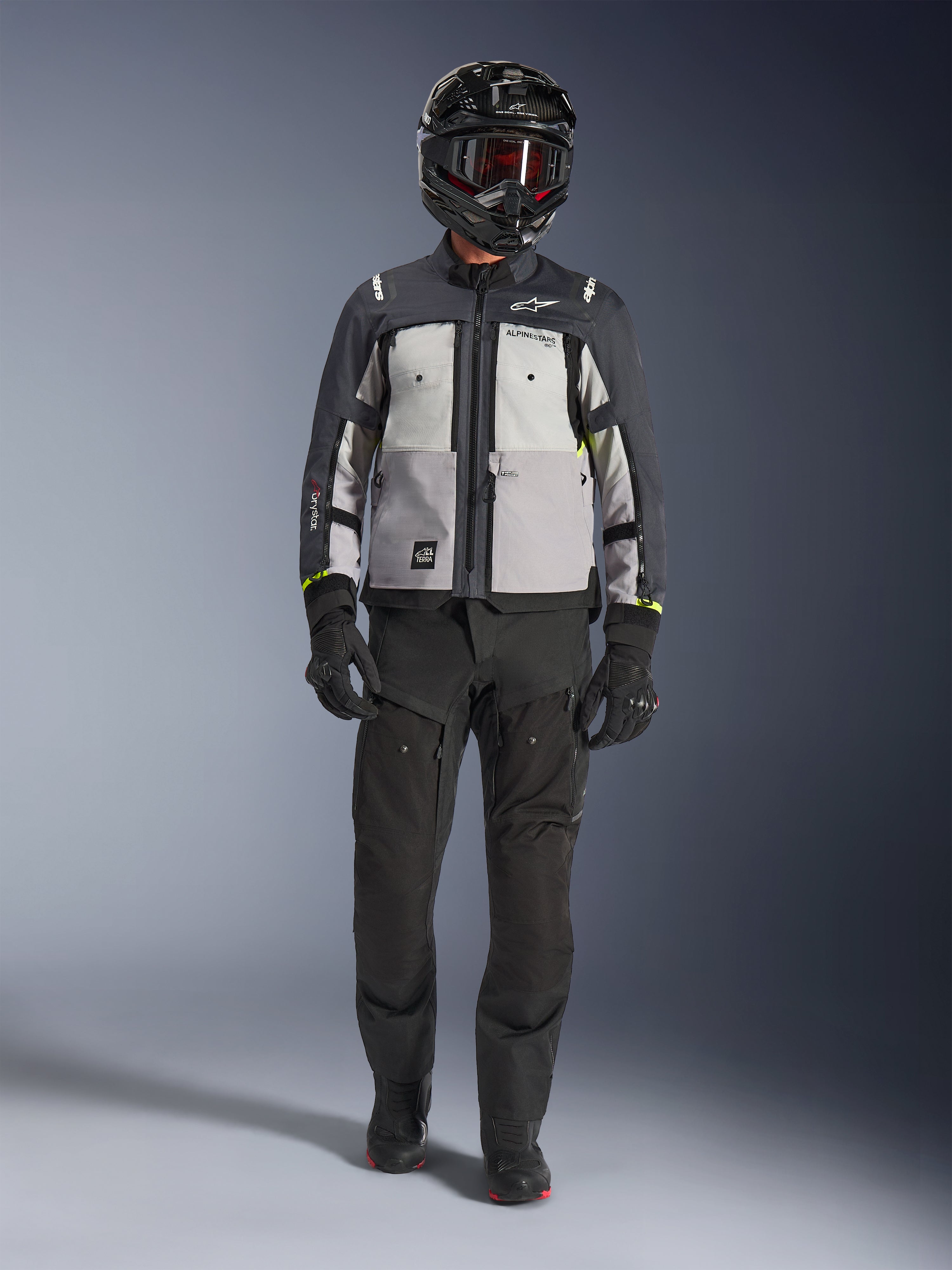 Chaqueta Alpinestars Cusco Drystar®, Ice Gray Mid Gray Deep Gray, chaqueta de motociclismo adventure combinada con pantalones de moto negros, guantes negros y casco negro con gafas, con paneles grises en bloques de color y detalles técnicos para touring y carretera