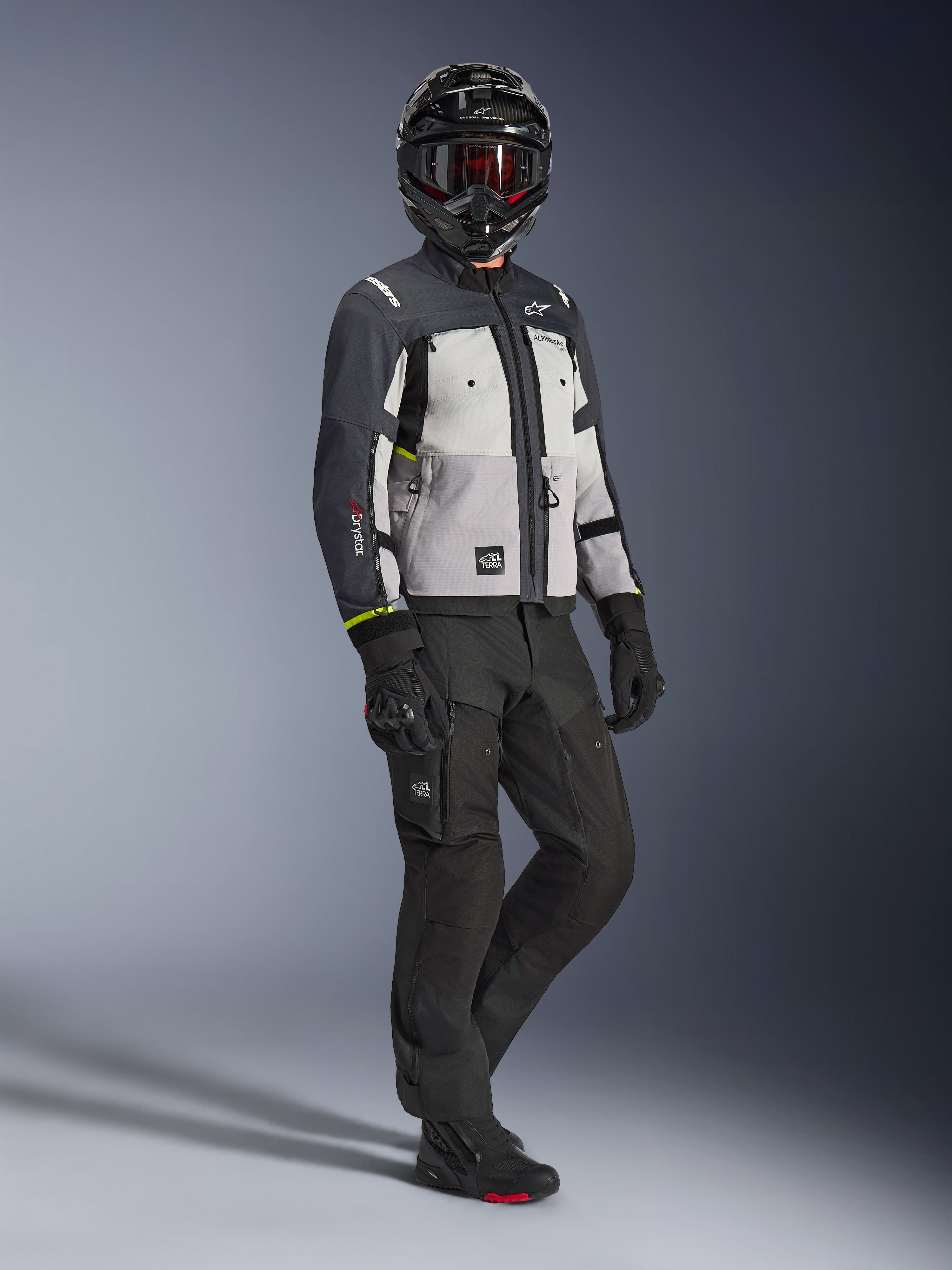 Chaqueta Alpinestars Cusco Drystar® en Ice Gray Mid Gray Deep Gray, toma de cuerpo completo de un motociclista con equipamiento de aventura completo que incluye casco negro con gafas rojas, pantalones negros y guantes a juego, chaqueta textil de moto con paneles de ventilación directa y membrana impermeable.