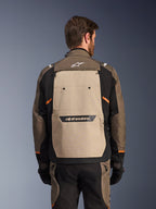 Chaqueta Alpinestars Cusco Drystar®, Walnut Aluminum Black, vista trasera, con un gran panel posterior color canela con logotipo negro, acentos naranjas, usada con pantalones y guantes a juego, diseñada para el turismo de aventura.