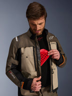 Chaqueta Alpinestars Cusco Drystar®, Walnut Aluminum Black, usada por un hombre que muestra el bolsillo para el protector de pecho con una inserción de armadura roja, chaqueta textil de motociclismo con ventilaciones con cremallera y logotipos para adventure touring.