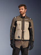 Chaqueta Alpinestars Cusco Drystar®, chaqueta de motociclismo adventure, nogal, aluminio y negro, usada por un modelo con guantes negros, con paneles frontales en color arena y mangas negras con logotipos de Alpinestars y parche All Terra, equipamiento de touring protector para todo clima