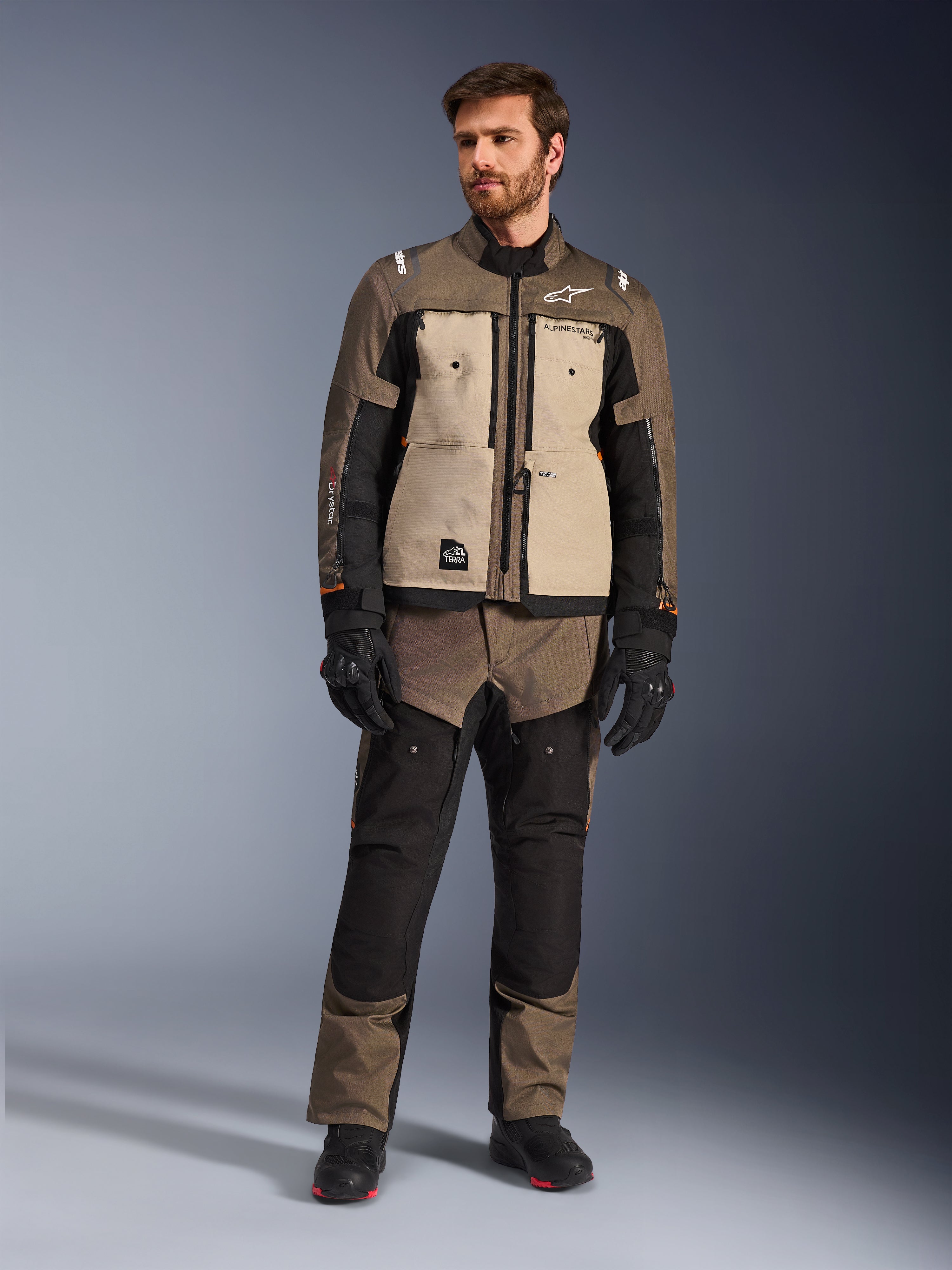 Chaqueta Alpinestars Cusco Drystar® en color Walnut Aluminum Black, vestida por un piloto masculino con pantalones de aventura touring a juego, guantes negros y botas, con paneles beige en el pecho, mangas negras con sutiles detalles en naranja y cremalleras de ventilación funcionales para la conducción en carretera.